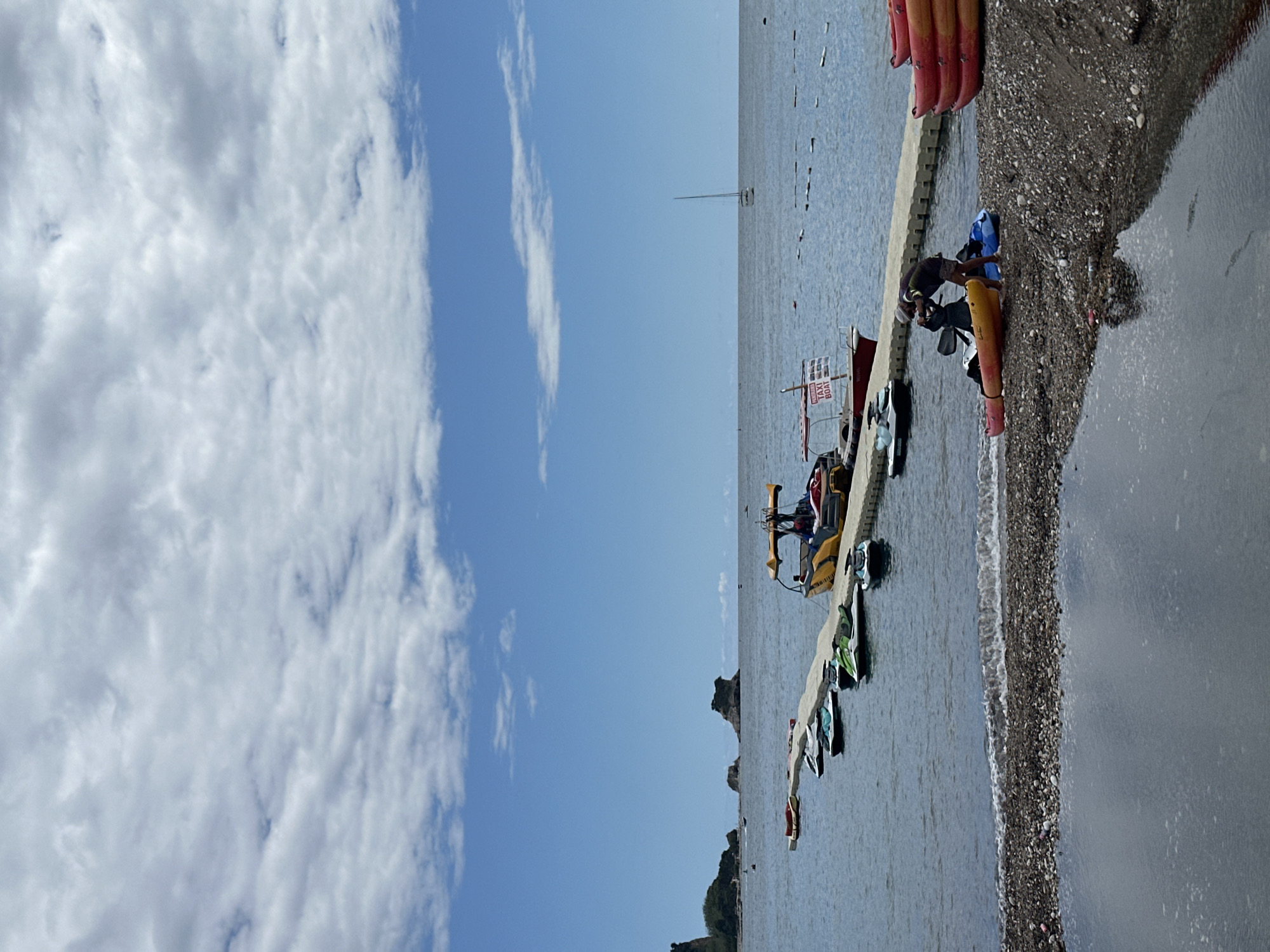 The image captures a sunny daytime scene at a coastal beach, offering a view of the sea, sky, and shoreline activities. The upper portion of the frame is dominated by a clear blue sky dotted with scattered white cumulus clouds.
Below the sky, a calm, light blue-grey sea extends towards the horizon. Along the right side of the water, a narrow, pebbly beach with gentle waves breaking on the shore is visible. A light-colored concrete pier or walkway stretches from the beach into the sea.
Various watercraft are present: several red and orange kayaks or paddleboards are stacked on the beach and along the pier. A series of colorful pedal boats, including yellow, green, and blue/white designs, are moored alongside the pier or floating in the water. One prominent yellow pedal boat is equipped with a slide. Further out on the water, a small boat with a mast can be seen near the horizon.
Several individuals are in the scene. One person in dark attire is on the pebbly beach near a blue kayak, seemingly engaged in preparing equipment or about to enter the water. Another person is visible on the yellow pedal boat with the slide. Near the pier, a small boat displays a red sign that clearly reads "TAXI BOAT" in white capital letters.
In the far distance, rocky islets or land formations are discernible, hinting at the geographical features of the coast. The overall impression is one of leisure and water-based recreation on a bright and pleasant day.