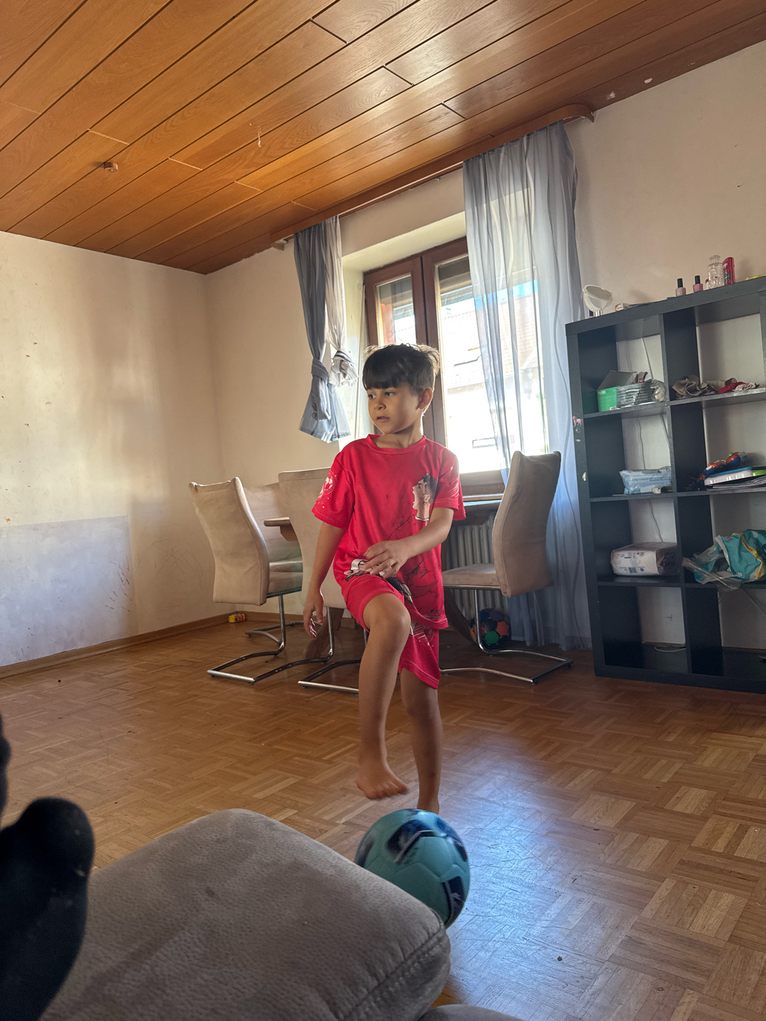 A young boy wearing a red t-shirt and red shorts is standing in a room with wooden floors and a wood-paneled ceiling. He is looking to the left, with his right leg slightly raised. A soccer ball is on the floor near his feet. In the background, there are chairs, a window with curtains, and a black shelving unit. The scene appears to be an indoor residential setting in Überherrn, Germany.