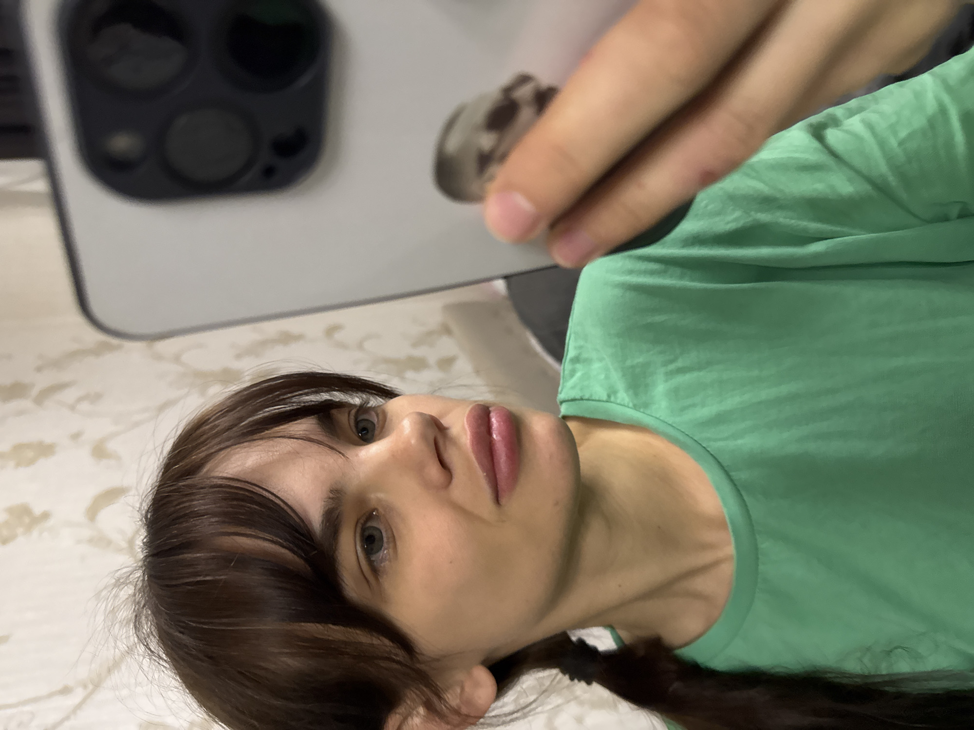 A person is visible in the foreground, angled to the left of the frame and positioned below the center. They have brown hair, light skin, and are wearing a green shirt. Above them, at the top of the frame, is a large, white object with a circular dark gray area that includes what appears to be camera lenses. A hand is holding the white object and there appears to be a small, dark object in between the finger tips. The background consists of a surface with a light-colored, patterned fabric. There is no indication of this image being taken in Rostov-on-Don, Russia.