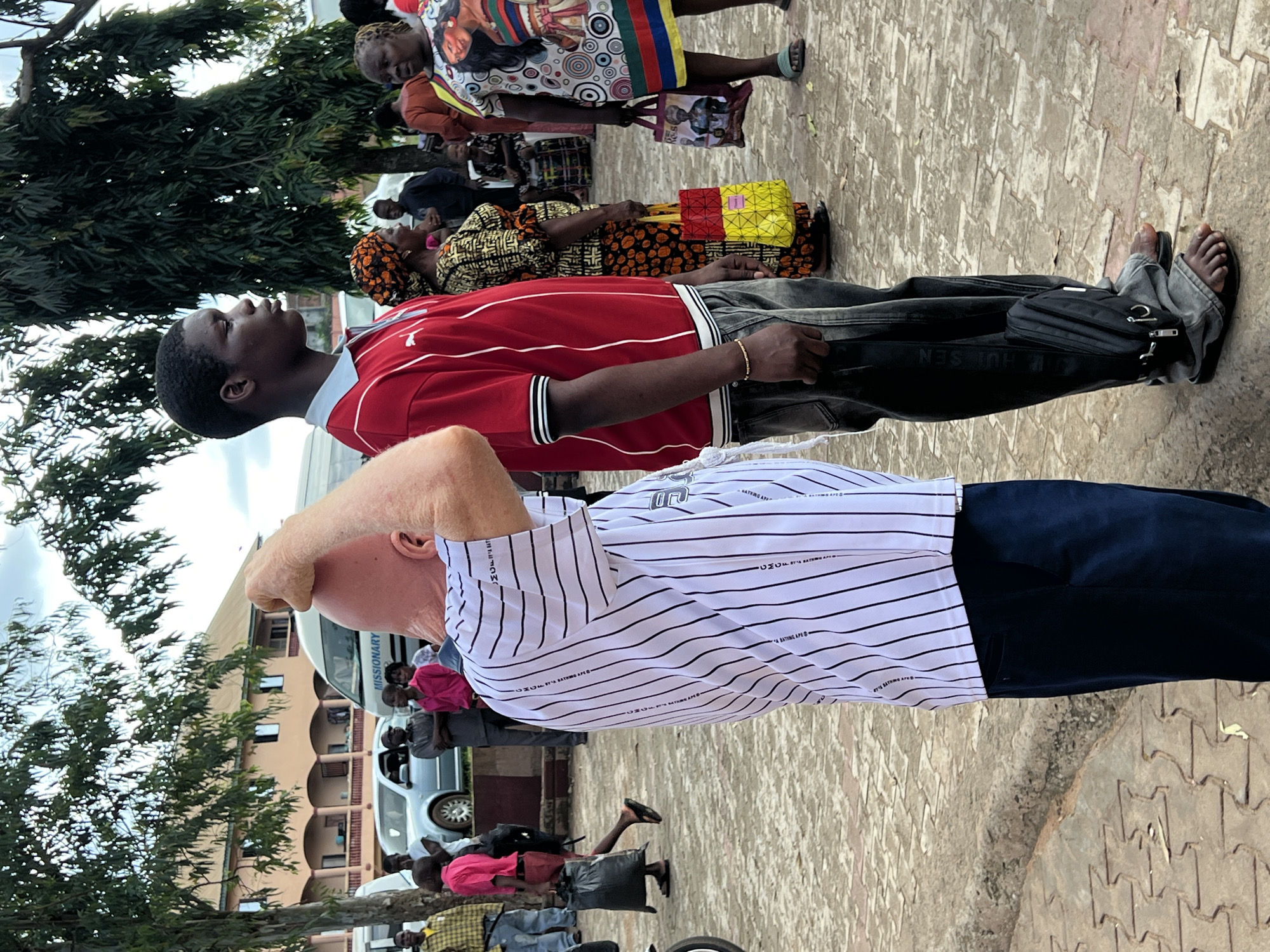 In Nnewi, Nigeria, a man with a shaved head, wearing a white and black striped shirt and navy blue pants, stands with his back to the camera. He is facing a group of people. To his right, a young Black man in a red polo shirt and dark pants looks away from the camera. Behind him, several people are visible, including a woman in a colorful patterned dress and another in a yellow and red patterned headscarf and dress, holding a shopping bag. Further in the background, a light-colored van with "MISSIONARY" written on it is parked near a building. The ground is paved with interlocking blocks.
