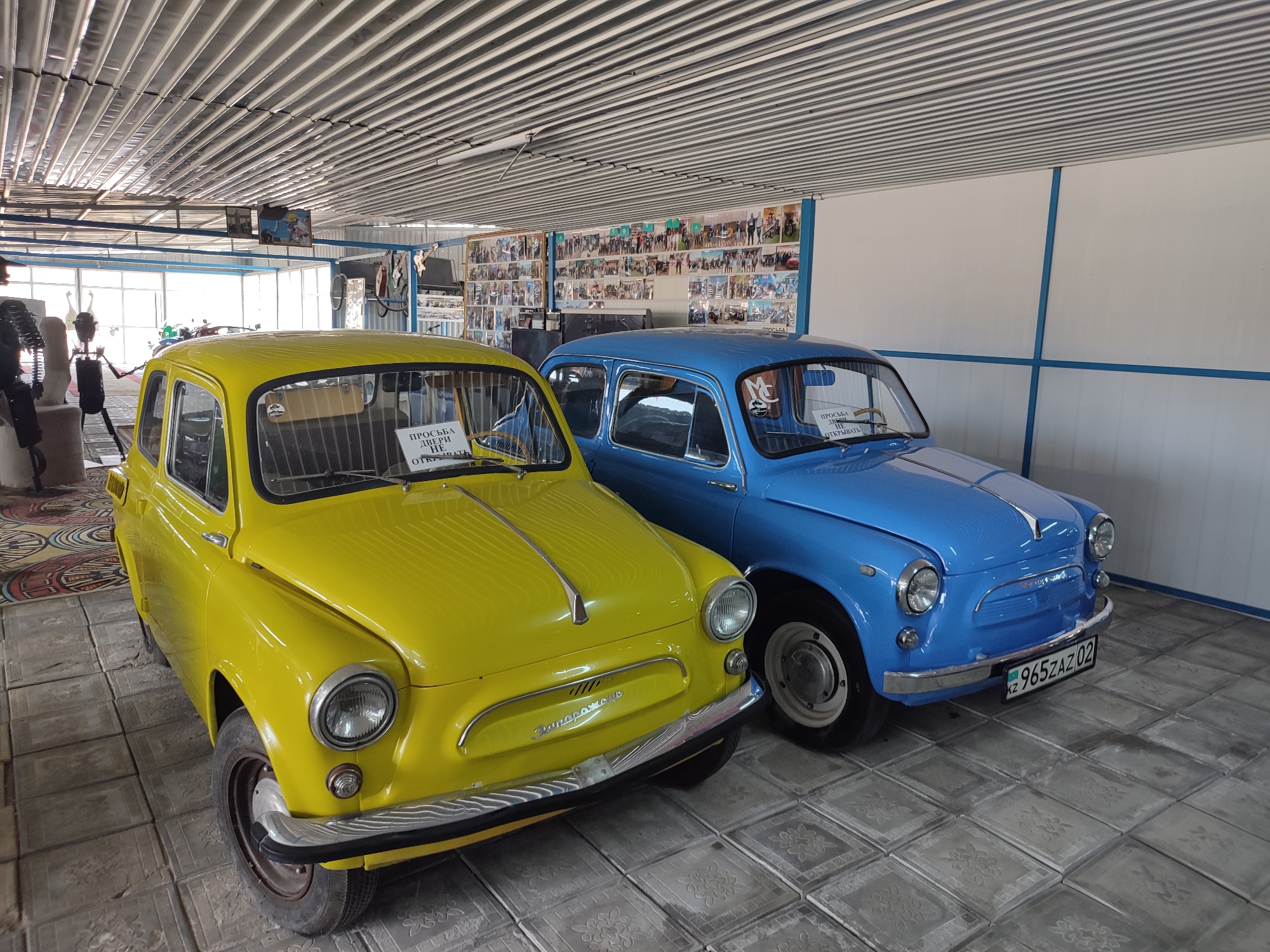 The image shows two vintage cars parked indoors. A bright yellow car is on the left, and a blue car is on the right. Both cars are small, older models. The yellow car has a sign on its windshield that reads "ПРОСЬБА ДВЕРИ НЕ ОТКРЫВАТЬ" which translates to "PLEASE DO NOT OPEN THE DOORS." The blue car also has a sign on its windshield. The license plate on the blue car is visible and appears to be a Kazakh license plate with the format "KZ 965ZAZ 02".
The cars are parked on a tiled floor with a floral pattern. The interior appears to be a museum or exhibition space, with a corrugated metal ceiling and walls adorned with many framed pictures. The overall impression is that of a display of classic vehicles.
Based on the license plate format, the location is identified as Qonaev, Kazakhstan.