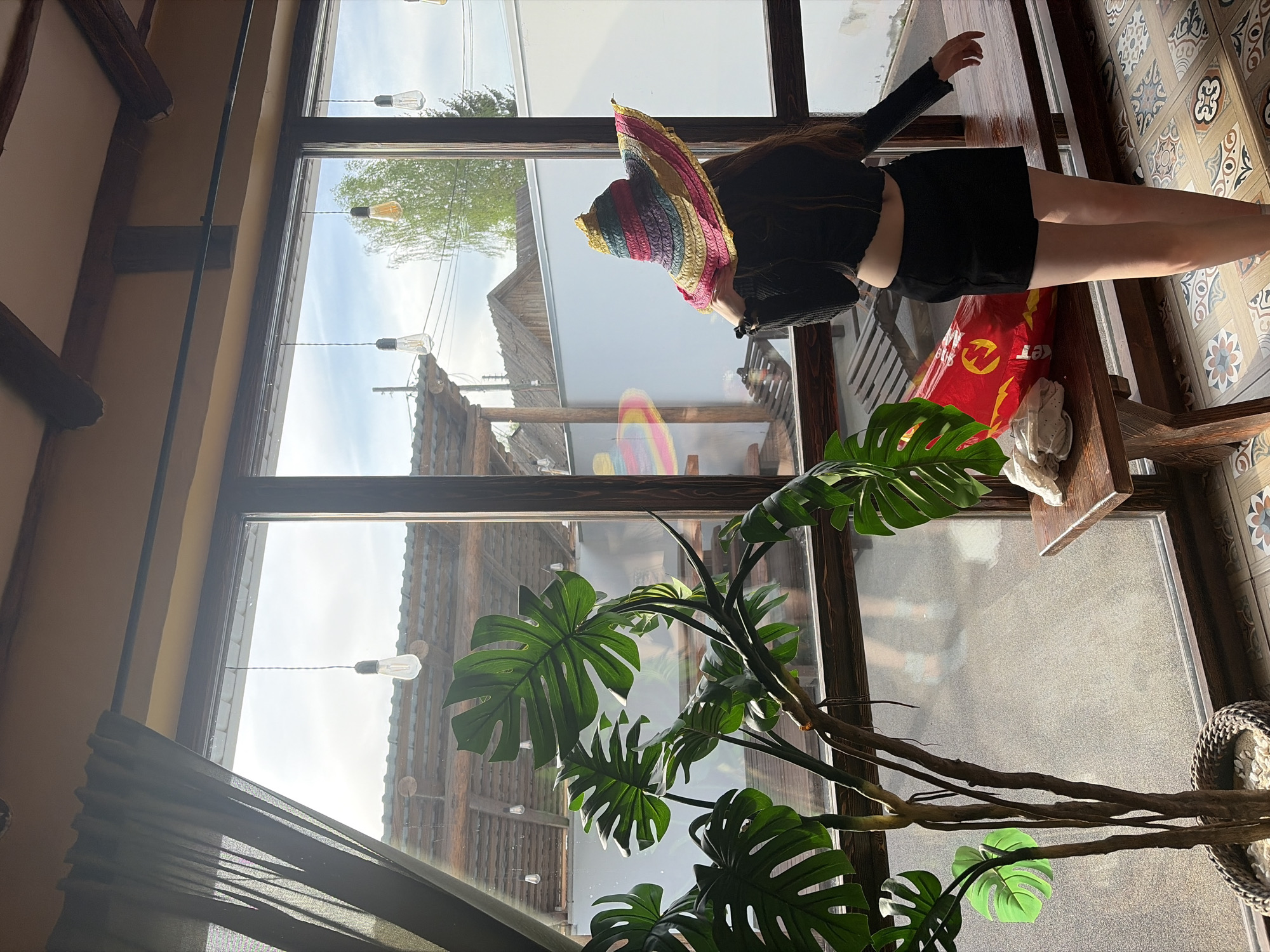 A woman in a colorful straw hat, black long-sleeved top, and black skirt sits on a wooden structure in front of a large window, possibly inside a cafe. The window shows a view of a village with traditional Russian buildings and hanging light bulbs. A large monstera plant occupies the foreground. The location may be Старое Михайловское, Russia.