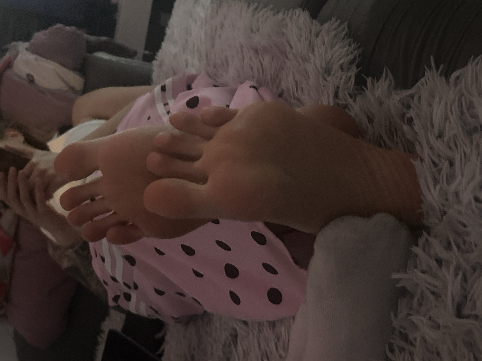 The image shows a person lying on a fluffy gray cushion with their feet prominently displayed in the foreground. The person is wearing a pink and black polka-dot top. One foot is partially covered by a gray sock. The other visible details include parts of the person's body, hands, and a blanket. The location in Burgas, Bulgaria, is unknown and cannot be determined from the image.