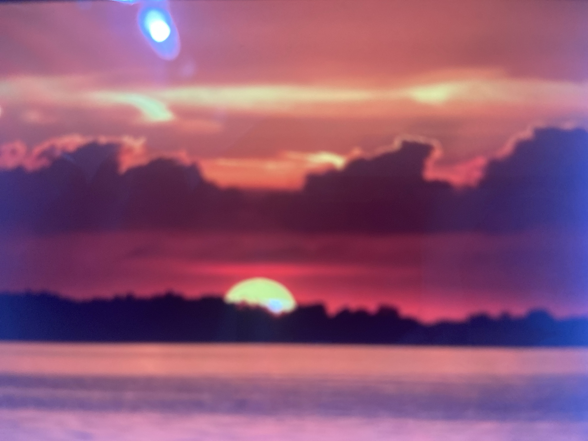 This is a blurry, horizontal, medium shot of a sunset over a body of water. The sky is a vibrant mix of pinks, oranges, and purples, with dramatic, dark clouds silhouetted against the colorful sky. A bright, pale yellow orb, likely the sun, is setting on the horizon, casting a soft glow. The foreground shows a dark, silhouetted treeline separating the water from the sky. The water itself is a calm expanse of muted pinks and purples, reflecting the colors of the sky. The overall mood is serene and tranquil, suggesting the end of a day. The image appears to be taken from a distance, possibly from across the water. There is no visible text in the image.