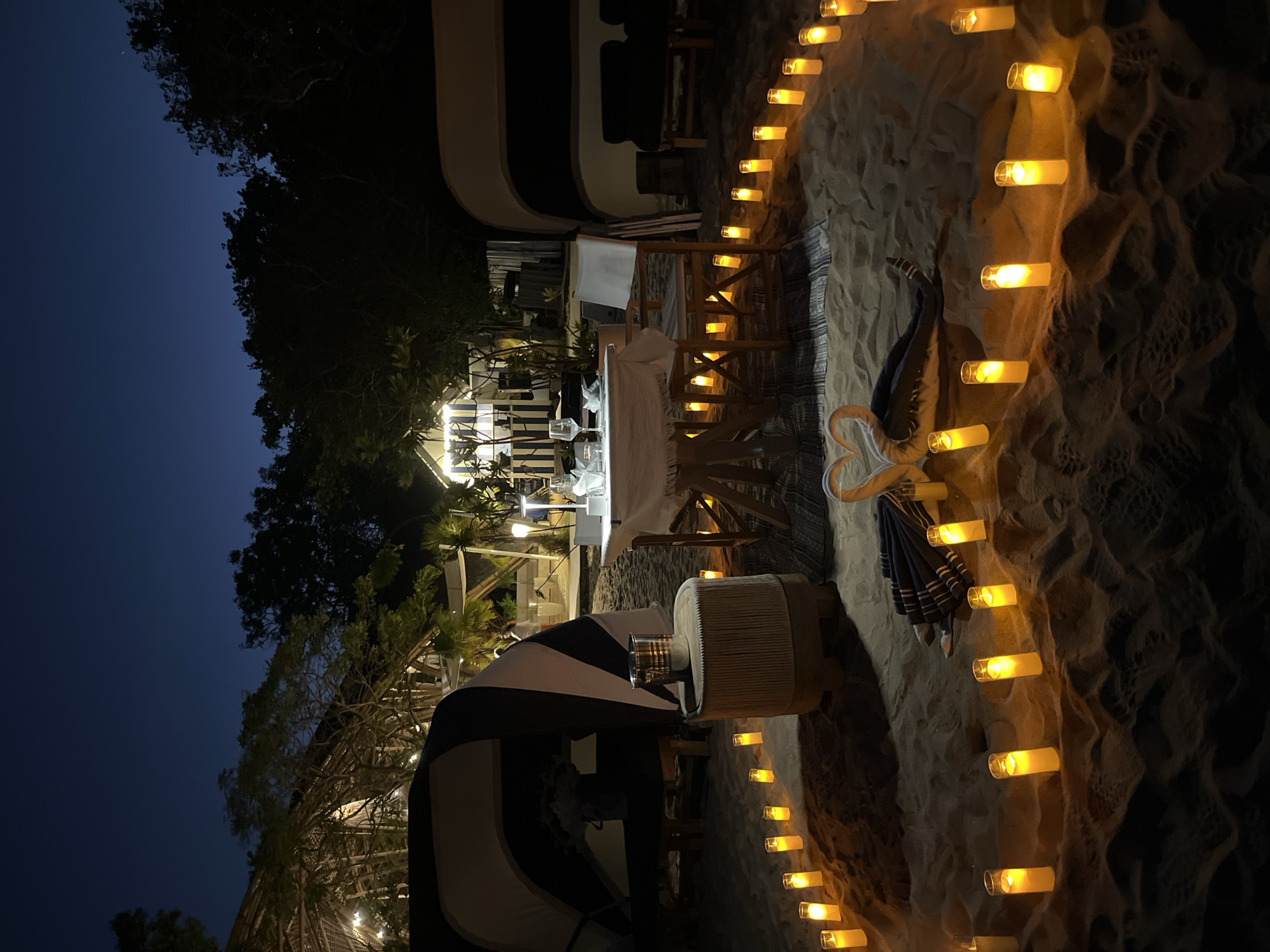 The image depicts an intimate, candlelit dinner setup on a sandy beach at night. The scene is located in Kuta Selatan, Indonesia, as evidenced by the surrounding lush vegetation and tropical architecture visible in the background.
In the foreground, a circular arrangement of pillar candles illuminates the sand, creating a warm and romantic ambiance. Towards the center of this circle, two white towels are folded into the shape of swans, placed beside a folded blue and white striped towel.
Further into the scene, a small, round table is set for a meal. It is covered with a white tablecloth and adorned with two wine glasses, cutlery, and a vase of flowers. Two chairs are positioned at the table, one with a white cushion. Adjacent to the table is a cylindrical stool with a textured surface.
Two large canvas tents, one on the left and one on the right, flank the dining area. They appear to be part of a glamping or luxury camping setup, with striped awnings. The tents and surrounding structures suggest a resort or private villa setting.
The background reveals dense foliage and trees characteristic of a tropical environment. Hints of structures with thatched roofs and modern, minimalist buildings with black and white stripes are visible, indicating a developed area within Kuta Selatan. The sky is a deep, dark blue, suggesting it is late evening or early night.
No people are directly visible within the immediate dining setup, though the presence of a resort suggests that staff or other guests might be present in the broader vicinity. The overall impression is one of a carefully curated romantic experience on a tropical beach.