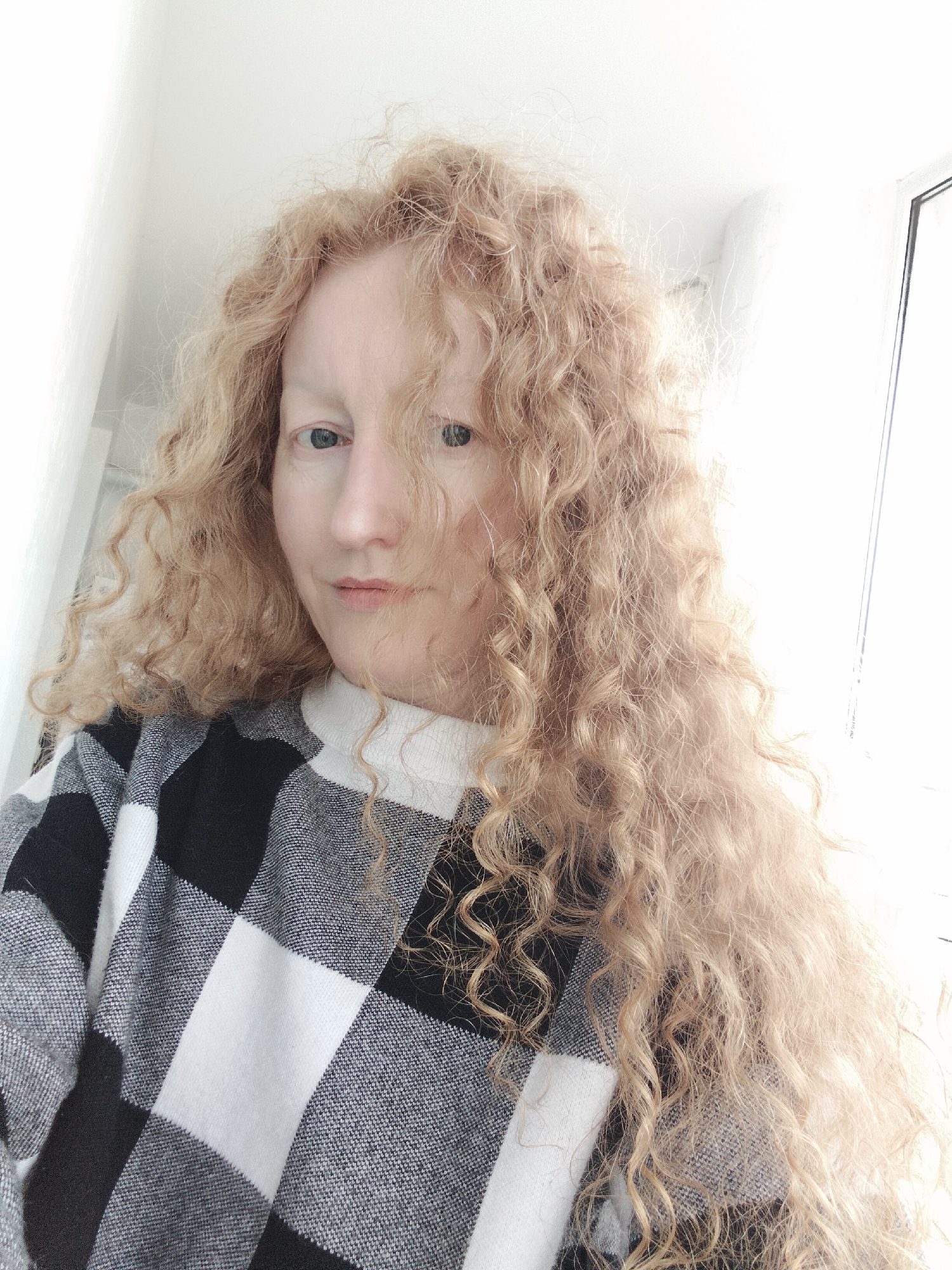A person with long, curly, light-ginger hair is centered in the frame, wearing a black and white checkered sweater. The individual has light skin, light eyebrows, and blue eyes. The background consists of a white wall and a bright window. The setting cannot be determined to be located in Krykhivtsi, Ukraine, without further information.