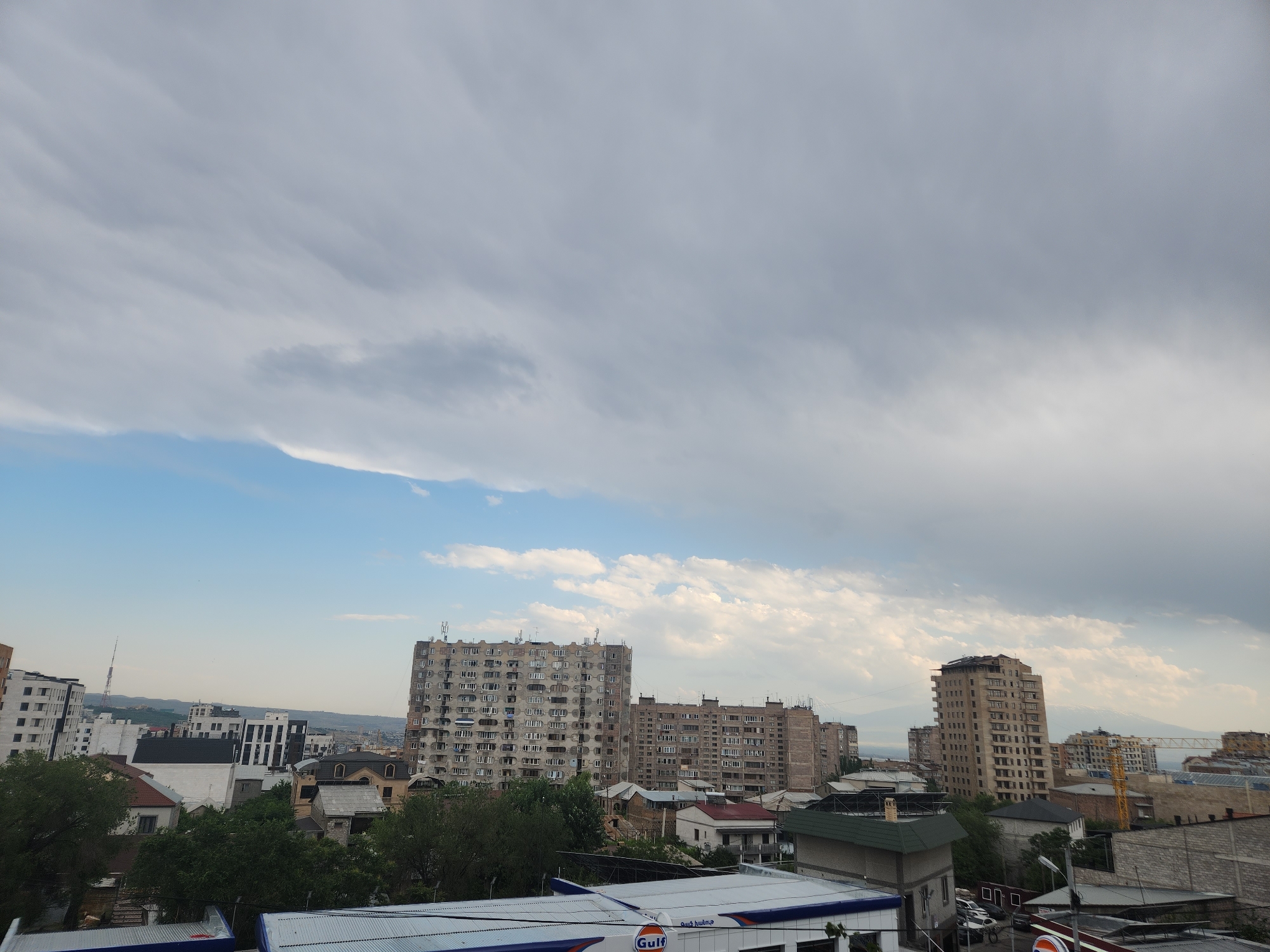 The image captures a panoramic view of an urban landscape under a partially cloudy sky. In the foreground, several rooftops are visible, one of which features a white canopy with a visible "Gulf" logo, indicating a gas station or a similar commercial establishment. Beyond this, a dense collection of buildings fills the midground, showcasing a mix of architectural styles. Dominating the center and right side of the frame are several multi-story apartment buildings, some appearing older with numerous balconies, while others are more modern with varied window designs. Smaller residential buildings and some trees are interspersed among the larger structures.
In the background, the cityscape extends towards a hazy horizon, with more buildings and some faint outlines of hills or mountains. The sky is a dynamic mix of bright blue patches and substantial grey clouds, with sunlight filtering through in some areas. The overall impression is of a densely populated city, possibly in a temperate climate, with a blend of old and new construction. The lighting suggests it might be late afternoon or early evening, given the diffused quality of the light and the dramatic cloud formations. No individuals are discernible in the image.