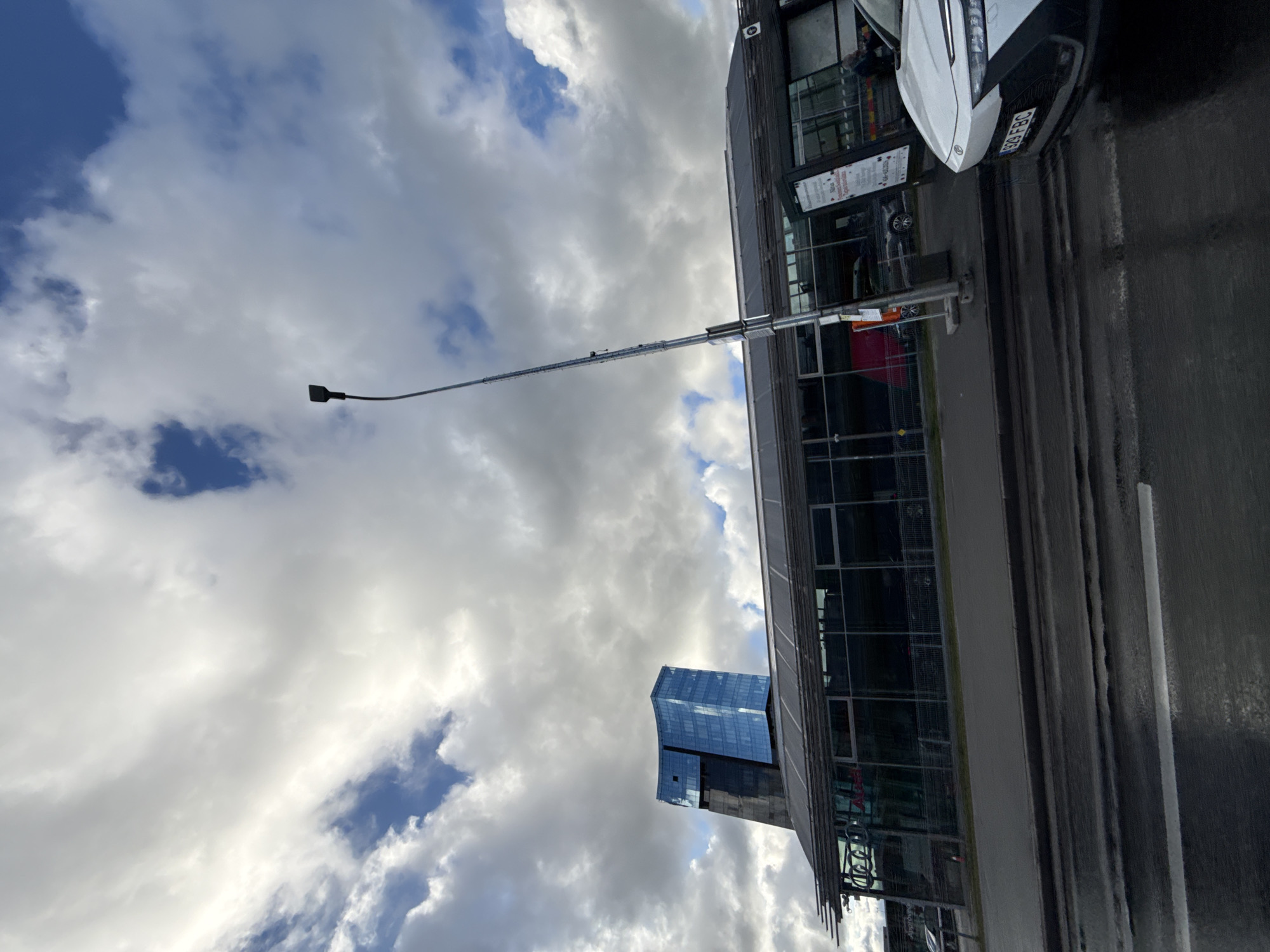 This image captures a view of the cityscape in Tallinn, Estonia, featuring a modern glass-fronted building and a sleek, white car. The building, characterized by its curved glass facade and dark, angular sections, stands prominently against a sky filled with dramatic, cloud-laden cumulus formations and patches of blue. In front of this building, a single-story structure with extensive glass panels and a metallic roof runs horizontally, possibly a commercial space or part of a transport hub. A tall, slender lamppost with a distinctive head extends upwards near the center of the frame.
Parked on the wet asphalt road in the foreground is a white, modern vehicle with a visible license plate reading "92G FBC". The road appears to have been recently wet, with reflections on its surface. The building's glass facade reveals reflections of the sky and clouds. Signs are visible on stands outside the single-story building. The overall impression is one of contemporary urban architecture contrasted with a dynamic, cloudy sky.