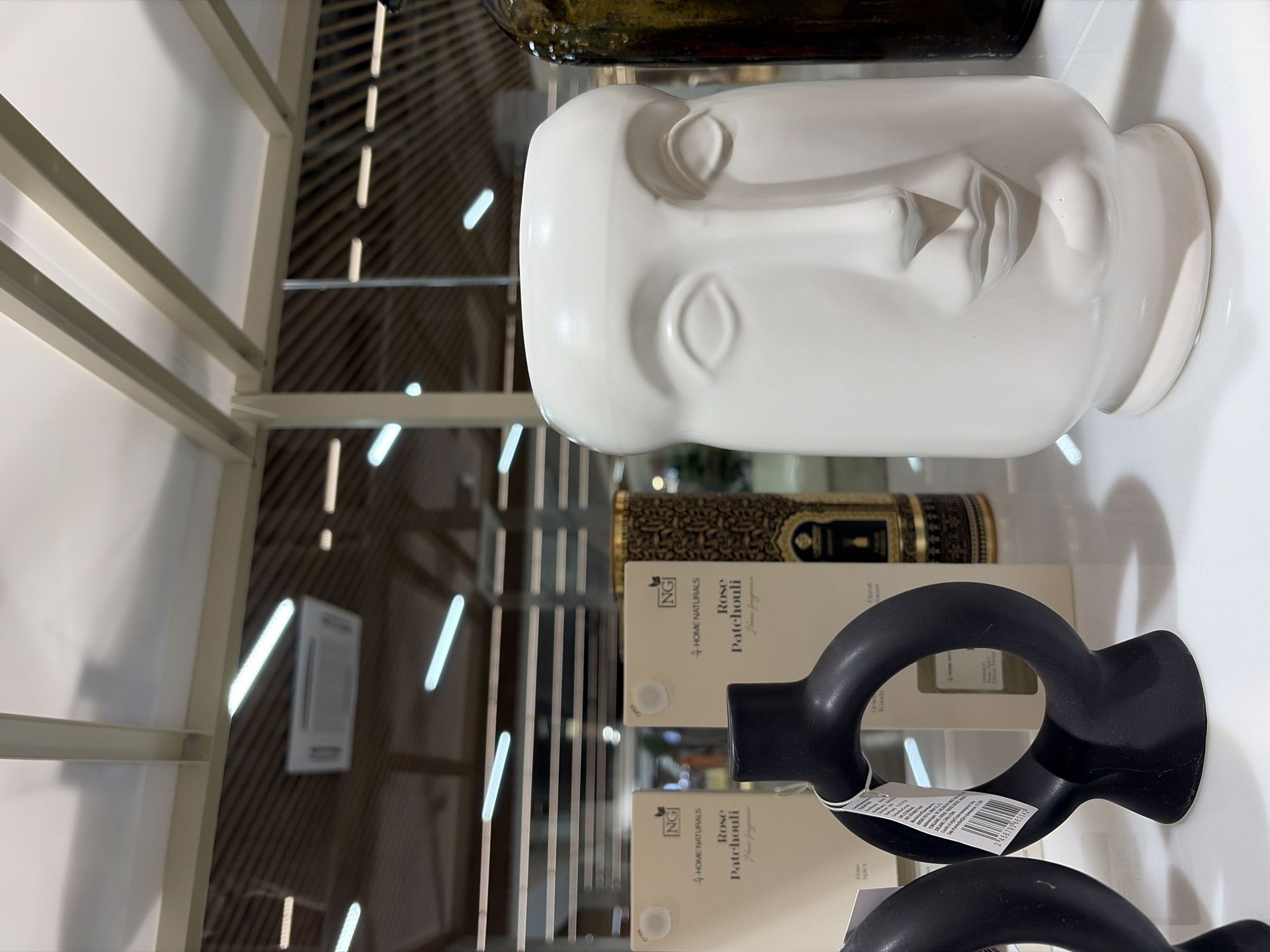 This indoor photograph, taken in Izhevsk, Russia, showcases a retail display of home decor items on a bright, reflective surface, likely a shelf or table in a modern home goods or gift shop.
The primary subject on the left is a white ceramic vase or sculpture shaped like a minimalist human face bust. It features simplified, hollowed-out eye sockets and subtly sculpted lips, resting on a small, flared base. Above and behind it, a portion of a dark, possibly green or brown, glass bottle is visible.
To the right, two black, matte-finished, abstract ring-shaped decorative objects are displayed, one in front of the other. These pieces have a smooth texture and a sculptural aesthetic. A white barcode sticker, with the number "2456790 662542," is attached to the front black object. Behind these items, several rectangular boxes are stacked. Visible text on the packaging includes "NG," "HOME NATURALS," and "Rose Patchouli Home fragrance." The word "OPEN" is also visible on one box, and "Wood" can be seen faintly on another.
The background reveals a modern interior with light-colored shelving components and a ceiling featuring linear fluorescent light fixtures, which are reflected in the shiny surfaces of the display. The scene suggests a clean, contemporary setting for showcasing decorative household items.