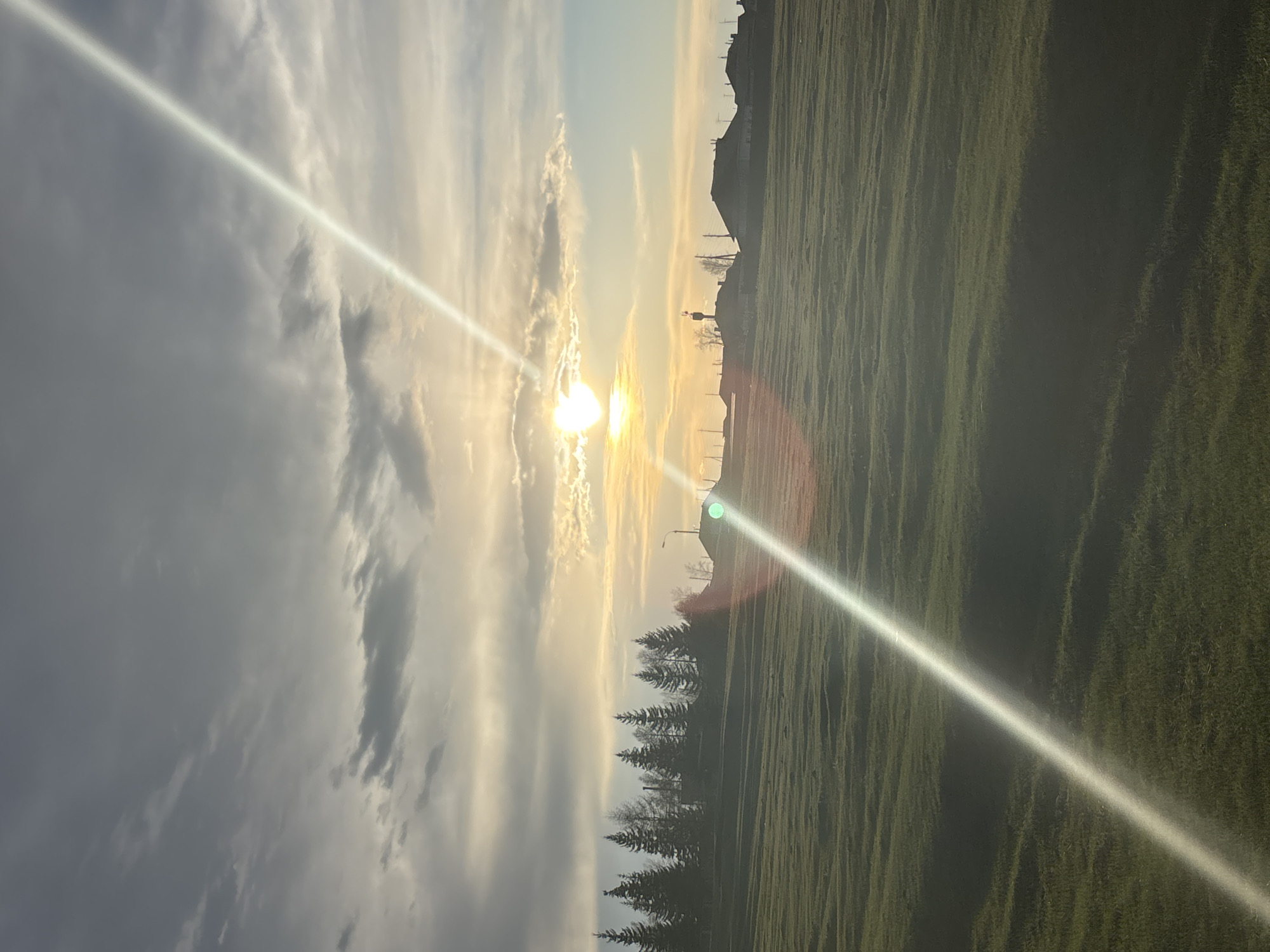 The image captures a landscape scene under bright sunlight, potentially near sunset or sunrise, in Zagatuy, Russia. The sun is visible through a break in the clouds, causing significant lens flare. The sky is filled with clouds of varying densities, ranging from dark gray to light, illuminated white clouds. The terrain consists of a grassy field, with trees bordering one section. In the background, silhouettes of buildings and what appear to be infrastructure, like a water tower, can be seen.
