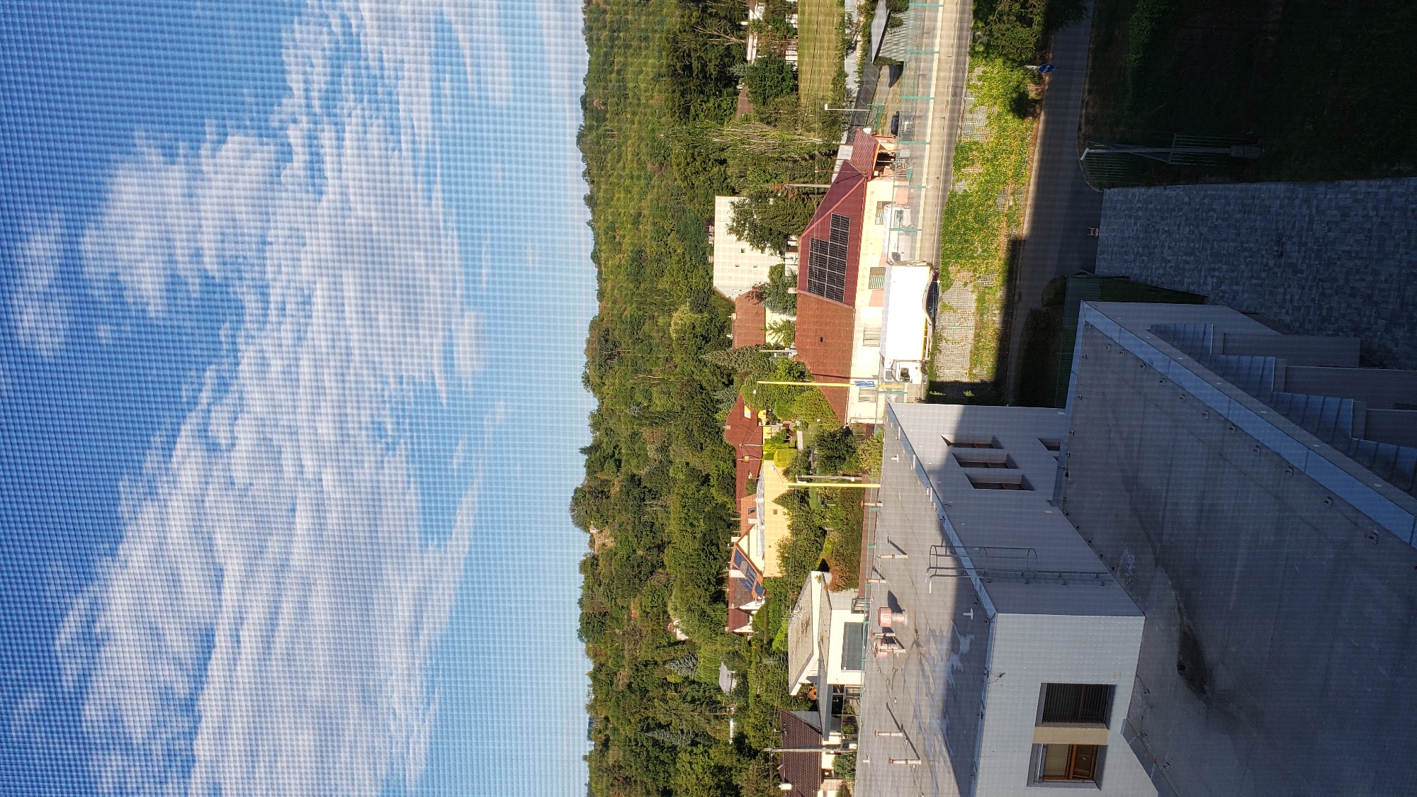 This image provides an elevated view of a suburban neighborhood in Prešov, Slovakia. The foreground features the rooftops of modern buildings, one with a flat, gray roof and another with visible ventilation pipes. A ladder is attached to the side of the building on the left. Beyond these buildings, a dense green hillside covered in trees dominates the background. Scattered among the trees are numerous residential houses with red-tiled roofs. Several of these houses are equipped with solar panels. A road runs horizontally across the middle ground, with a white truck and a few cars visible. A green metal fence separates the road from a grassy area with some vegetation growing on a textured wall. The sky is blue with scattered white clouds, indicating daytime. The scene appears to be taken from a high vantage point, possibly a building or a hill overlooking the neighborhood.