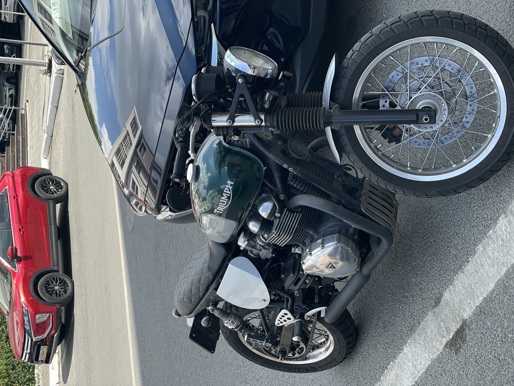 A green Triumph motorcycle is parked on asphalt next to a black car. A red Mitsubishi Eclipse Cross SUV is parked in the background. The motorcycle has a quilted seat, chrome accents, and a black exhaust pipe. The license plate of the red SUV reads "E540KM716". The scene appears to be a street in Kazan, Russia, as indicated by the license plate format typical for Russia and the context.