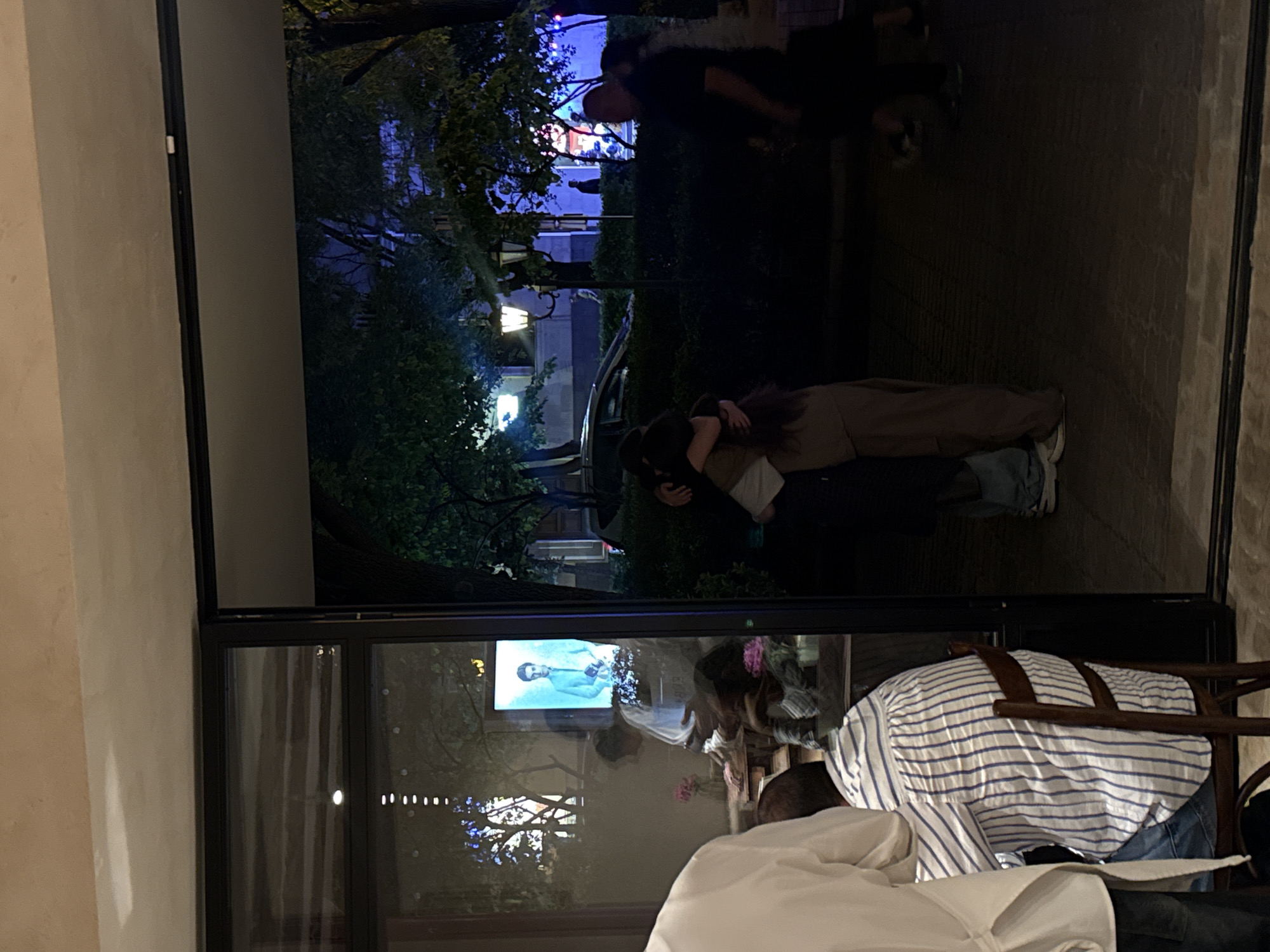 Here is a detailed description of the image:
**Location:** The image appears to be taken from inside a restaurant or bar in Yerevan, Armenia, looking out through a large window or glass door. This is inferred from the objects inside (tables, chairs, people) and the outdoor scene visible beyond.
**People:**
* **Inside:** Several people are partially visible inside the establishment. A person in a striped shirt is seated in a wooden chair. A person in a white shirt is also visible. The partial reflections in the windows suggest other people are present.
* **Outside:** A couple is hugging on the street outside. Another group of people is walking down the street.
**Subjects:**
* **Exterior:** The outside scene features trees, street lamps, a paved area, and a building in the background illuminated with blue lights.
* **Interior:** The interior shows elements of a restaurant or bar setting.
**Objects:**
* Tables, chairs, wooden chair, window or glass door, clothing, street lamps, building.
* There is a television screen reflecting the outdoor scene.
**Actions:**
* People hugging, people walking, dining.
**Interactions:**
* People embrace outside.
* People are dining inside.
**Environment:**
* The time of day is likely evening or night, given the lighting.
* Outside, there is an urban environment with trees and buildings.
* The restaurant/bar has a modern design, inferred from the large windows and visible interior details.