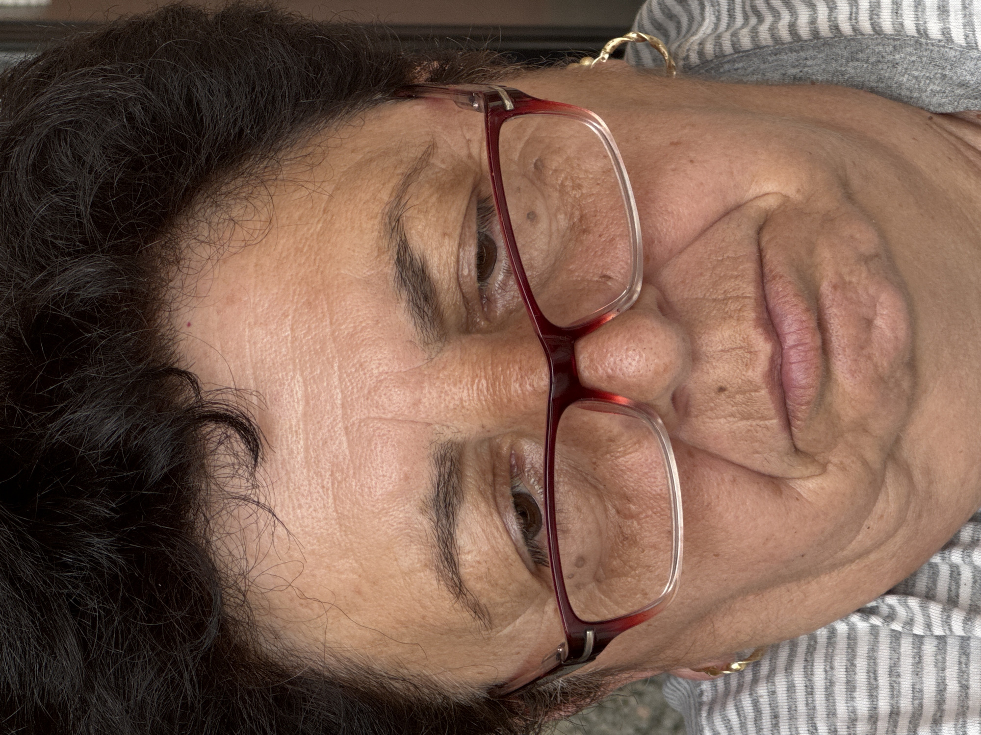 A medium shot of an older woman with dark, curly hair wearing red-framed glasses and a striped shirt. Her skin is fair with visible wrinkles, and she is looking downwards. The context of Okop, Bulgaria is not apparent from the image itself.