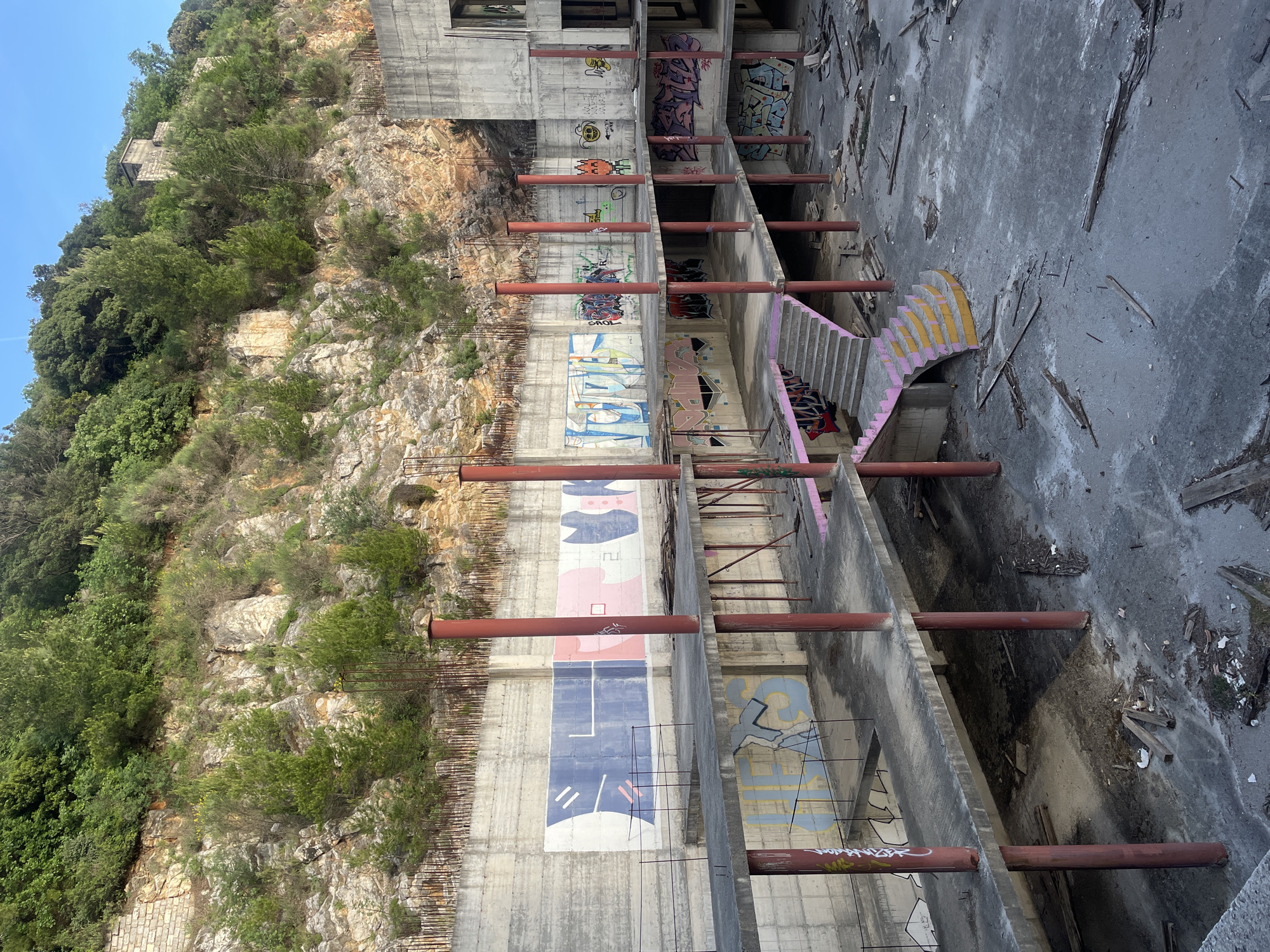 The image depicts the ruins of the abandoned Hotel As in Donji Katun, Montenegro. The multi-story concrete structure is built into a rocky hillside covered with vegetation. The hotel's exposed framework reveals graffiti and faded artwork on the interior walls. Red support columns extend horizontally across the open floors. A staircase with pink and yellow painted edges connects some levels. The interior floor is littered with debris, including pieces of wood. The sky is visible at the top of the frame.
