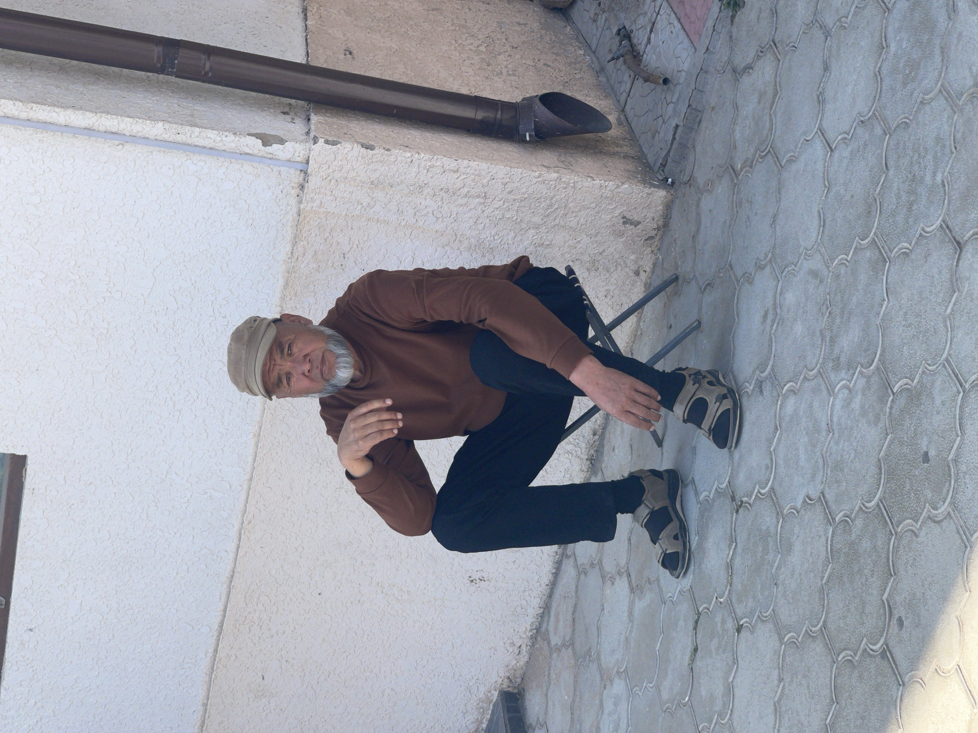 An adult male is sitting on a small, portable, dark gray stool on a paved area. He is wearing a light brown cap, a long-sleeved brown shirt, black pants, and gray sandals with black straps. He has a gray beard and mustache. His left arm is resting on his knee, and his right hand is on his lap. Behind him, a textured off-white wall runs horizontally, with a dark brown downspout attached to the right side of the wall. The ground is paved with interlocking light gray tiles. The scene appears to be outdoors, possibly in a courtyard or alleyway. The overall lighting suggests it is daytime.