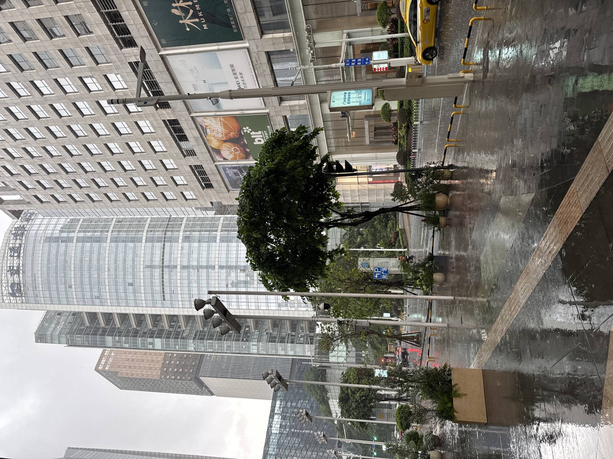 This is a medium shot of a street scene in Tianhe District, China, on a rainy day. The foreground is dominated by a wet, dark-tiled sidewalk, reflecting the surrounding buildings and streetlights. A tactile paving strip runs diagonally across the sidewalk. Several potted plants and planters are placed along the edge of the sidewalk. A yellow taxi is visible in the middle ground, driving on the wet asphalt road, which also shows reflections. In the background, tall, modern buildings with glass facades and concrete exteriors rise into the overcast sky. Streetlights and traffic signals are present, with the signals indicating green and red lights. The weather is inclement, with visible rain. No people are clearly identifiable in the scene. The overall impression is one of a busy urban environment experiencing a downpour.