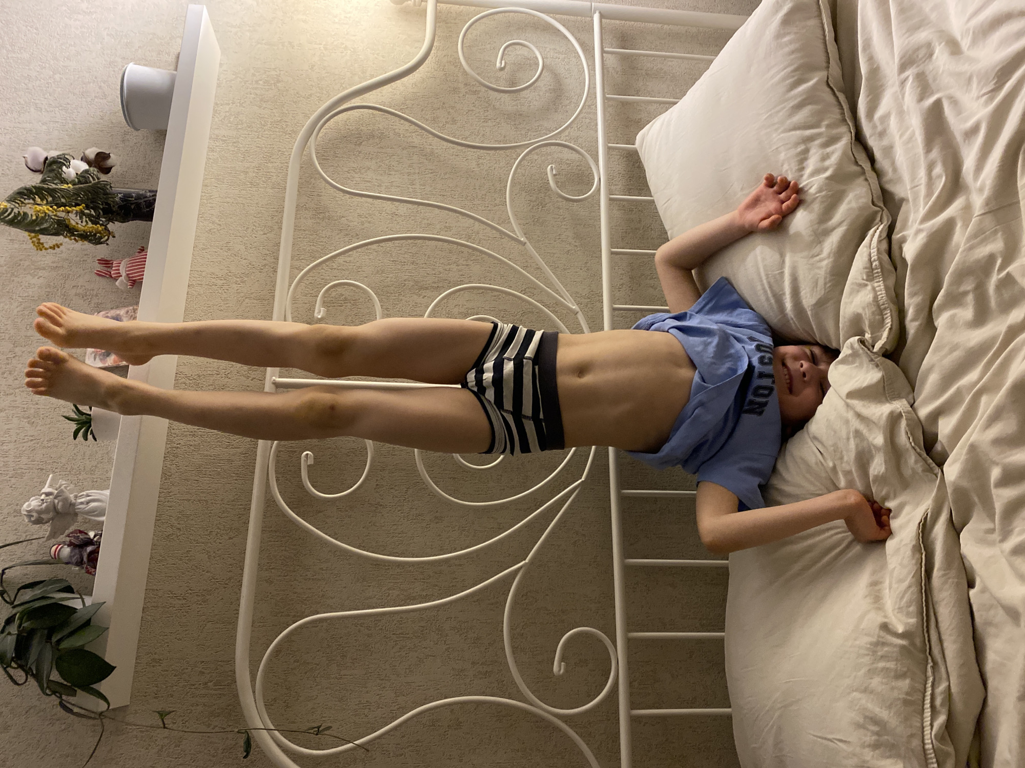 A child is lying on a bed in a bedroom. The child is wearing a blue t-shirt and striped underwear. The bed has a white metal frame. There is a shelf on the wall with plants and other decorative items. The room is lit by artificial light. The location is not discernible from the image.