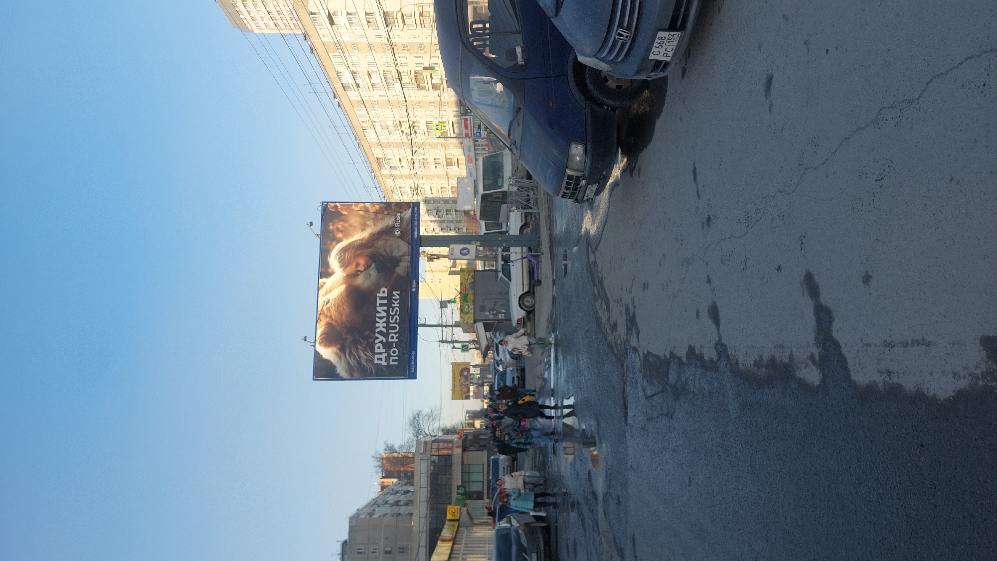 The image depicts a street scene in Novosibirsk, Russia. A large billboard dominates the left side, featuring an advertisement with a woman and a dog, with the Russian text "ДРУЖИТЬ ПО-RUSSKI". Below the billboard is a street with a mix of people walking and standing. A variety of vehicles are present, including cars, vans, and trucks. The street appears wet, possibly from rain or washing. Buildings are visible in the background, including residential structures. A blue car with a license plate is partially visible at the top right of the image. Overhead wires run across the sky.