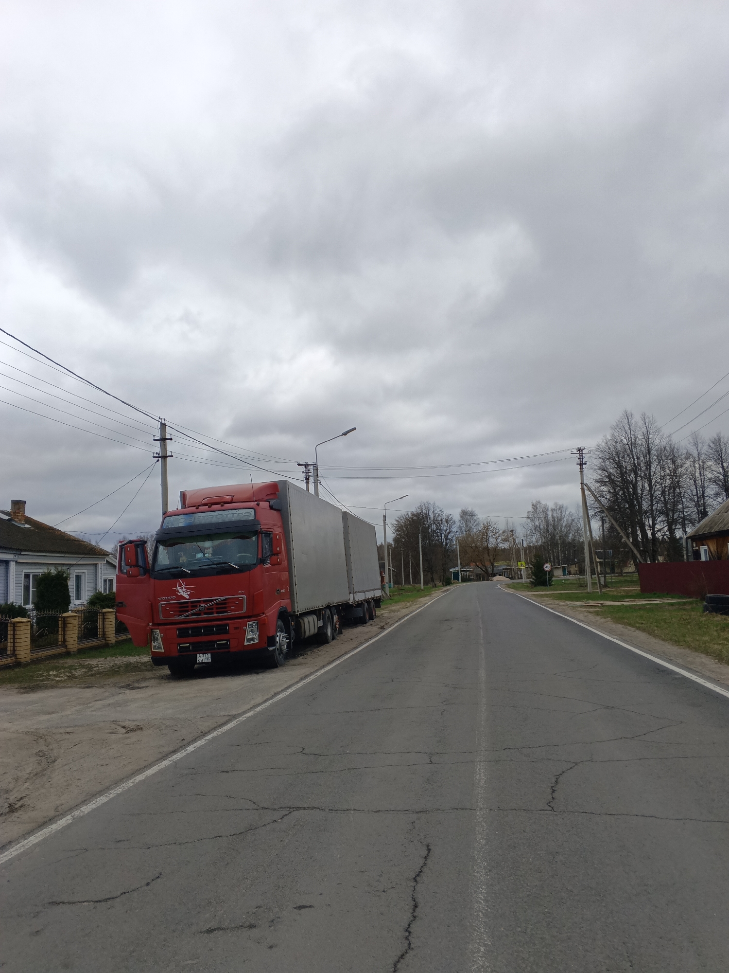 The image depicts a red Volvo FH truck parked on the side of a cracked asphalt road in Красный Маяк, Russia. The truck's driver-side door is open. Power lines are visible overhead, supported by utility poles on both sides of the road. Buildings are visible on either side of the road, along with bare trees. The sky is overcast.