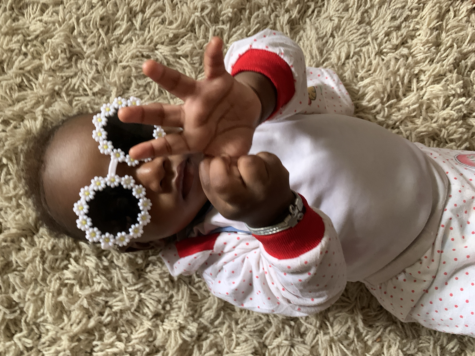 A baby lies on a beige shag carpet. The baby wears a white long-sleeved shirt with red polka dots and red cuffs, and round, white sunglasses decorated with small, daisy-like flowers. The baby's right hand is raised with fingers spread, while the left hand is in a fist near the face. A silver bracelet is visible on the baby's left wrist. There is no indication that this image was taken in Barakin Akawo, Nigeria, so that location cannot be determined forensically.