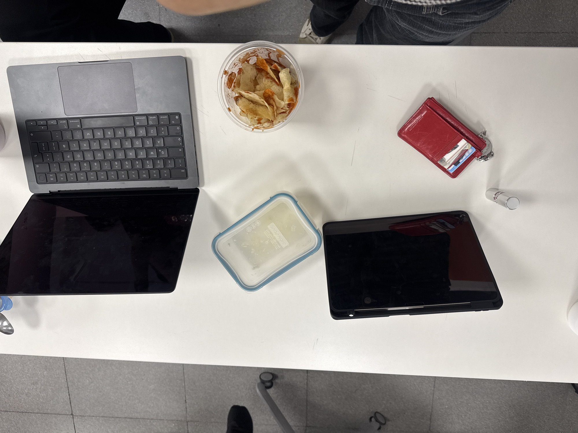 The scene depicts a white table surface with several items arranged on it, seemingly in an office or study area. On the left side is a partially opened gray laptop with a visible keyboard and screen. Next to it is a clear plastic container filled with what appears to be food, possibly chips or snacks. Towards the right side of the table, there is a red wallet or cardholder with cards visible inside, along with a small silver tube, possibly lipstick or a similar cosmetic product. Below these items, there is a rectangular plastic food container with a blue rim, next to a black tablet, possibly an iPad. The scene is lit well with overhead lighting and shows parts of people's clothing on the periphery; the top of a man's trousers and legs on top left, and the top of a person's legs on the top right. The floor is visible in the bottom portion of the image and appears to be tiled with a gray pattern. The scene appears to be a candid snapshot of someone's workspace.
There is no information about this photo being in the city of Puebla, Mexico.