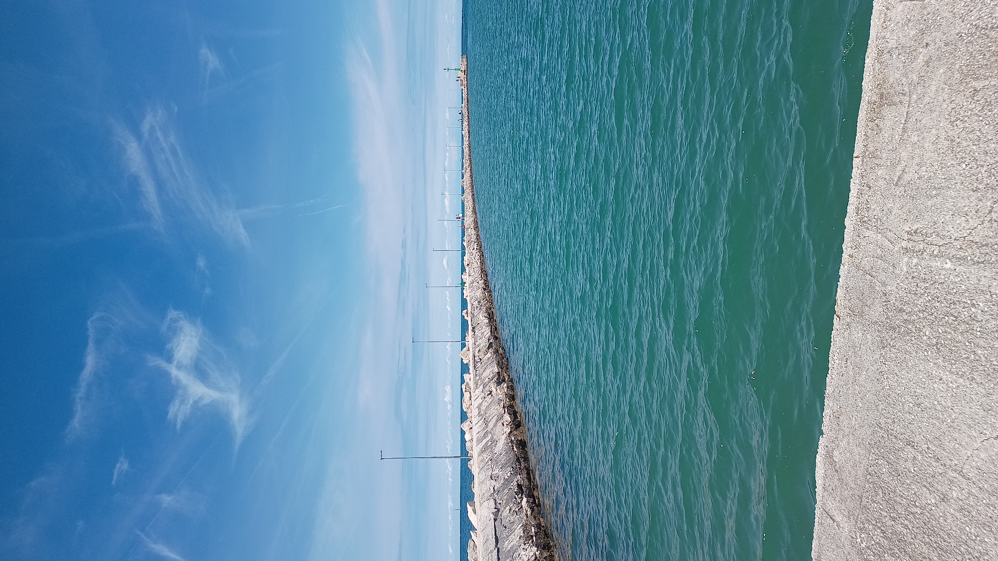 This media file presents an expansive coastal scene featuring a long pier extending into the sea under a bright sky. The image is oriented vertically, but when viewed horizontally, it depicts a clear horizon separating the blue sky from the vast expanse of turquoise-blue water.
In the upper half, the sky is a brilliant, clear blue, adorned with delicate, wispy white clouds, possibly cirrus formations or faint contrails from aircraft, suggesting fair weather. The lower half of the image is dominated by the calm, rippling surface of the sea, which transitions from a deeper blue-green closer to the foreground to a lighter hue towards the horizon.
A prominent concrete pier or breakwater stretches from the foreground into the distance, delineating the sea from a walking path. The pier's edge facing the water is lined with rugged, light-colored rocks. Along the pier, a series of slender, dark poles, likely lighting fixtures or bollards, are evenly spaced, receding into the background. In the far distance, at the very end of the pier, a small red and green structure, possibly a navigational beacon or a miniature lighthouse, is visible. A few indistinct figures, presumably people, can be faintly seen walking on the pier further down.
The foreground on the right features a section of the concrete walkway, exhibiting a rough, textured surface. The time of day is clearly daytime, characterized by bright, even sunlight. The overall atmosphere is peaceful and serene, showcasing the natural beauty of the coastline. This scene is identified as being in Umag, Croatia. No visible text is present in the image.