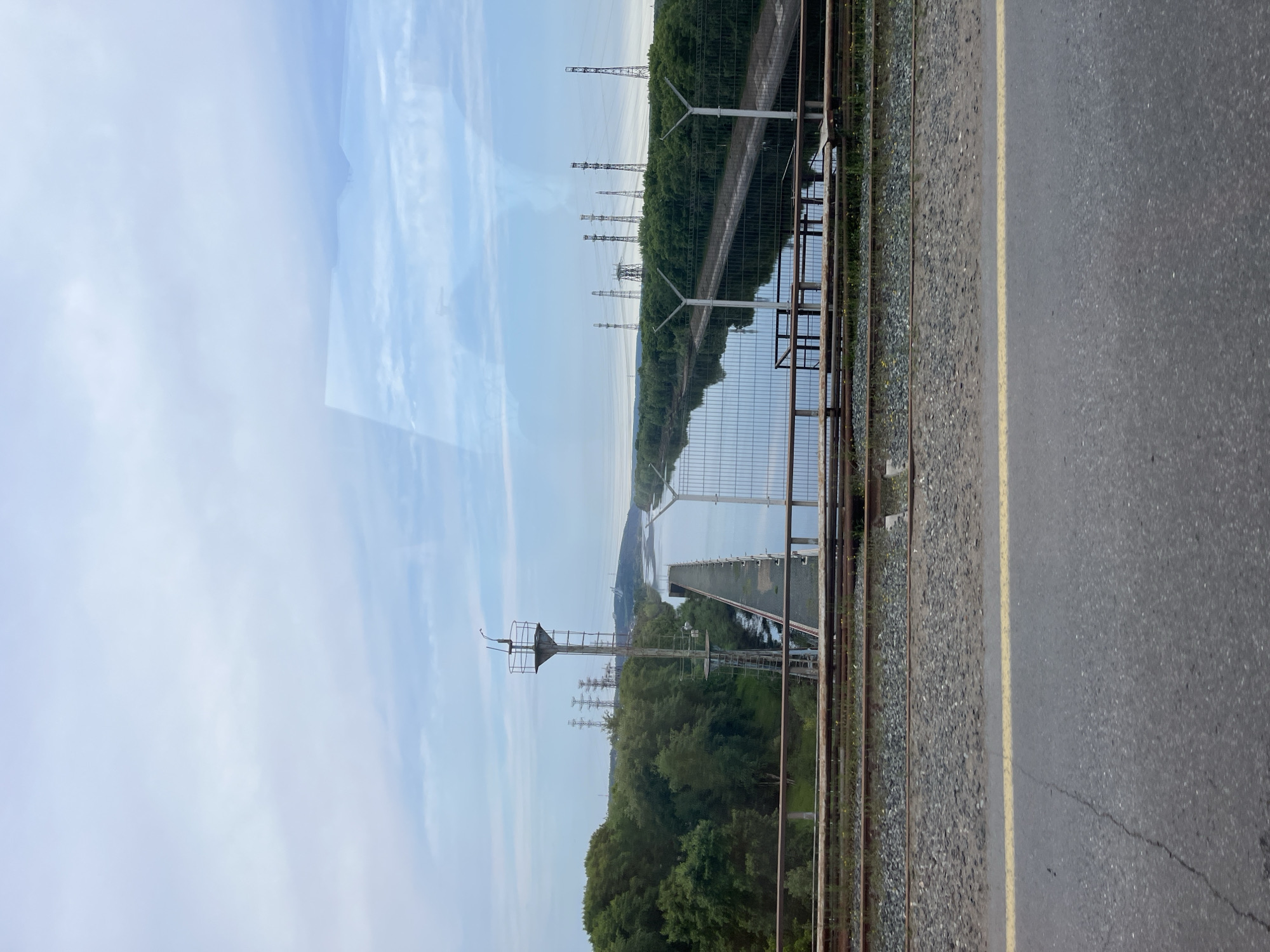 The image presents an outdoor landscape featuring a blend of natural scenery and human-built infrastructure, likely located in Chaikovsky, Russia. The perspective is from a slightly elevated vantage point, looking across a body of water.
In the immediate foreground, an asphalt road with a solid yellow dividing line runs parallel to a set of railway tracks, which are laid on a bed of gravel. A sturdy metal fence, composed of vertical posts and horizontal bars, separates the railway tracks from the area beyond.
Immediately past the fence, a wide expanse of water, appearing to be a river or a large canal, stretches into the distance. On the opposite bank of this waterway, a dense band of green trees forms a forested area that extends along the horizon. Several tall, metallic electricity transmission pylons rise above the treeline, spaced out and continuing into the far distance, suggesting a power line corridor. Closer to the viewer, positioned near the railway tracks, stands a prominent, tall metal signal or communication tower, equipped with platforms near its apex. A faint, bridge-like structure is also visible crossing the water further in the background.
The sky is a pale blue with scattered, wispy white clouds, indicating a clear or partly cloudy day. The soft, even lighting suggests it is either morning or late afternoon. There are no people visible in the scene, and no discernible text can be found within the image.