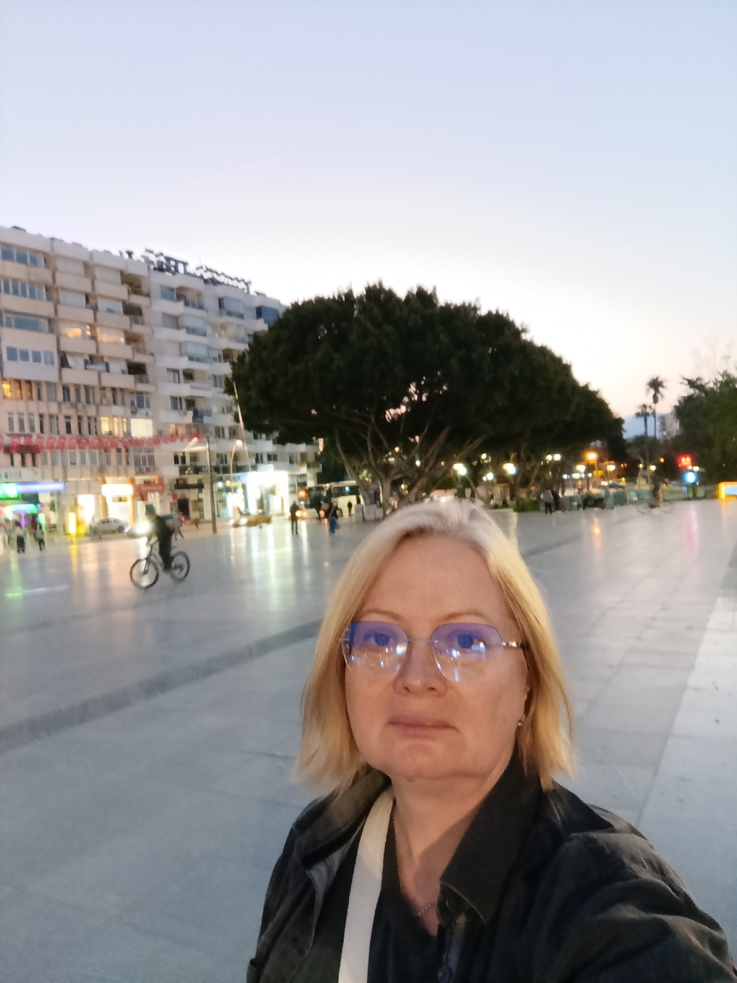 A woman with blonde hair and glasses takes a selfie in a city square at dusk. She is wearing a dark jacket over a light-colored top. Behind her is a paved square with people walking and a person riding a bicycle. There are several lit trees and street lights. In the background is a multi-story building with lights on. The image appears to be taken in Antalya, Turkey, based on the style of architecture and urban design of the buildings and square.