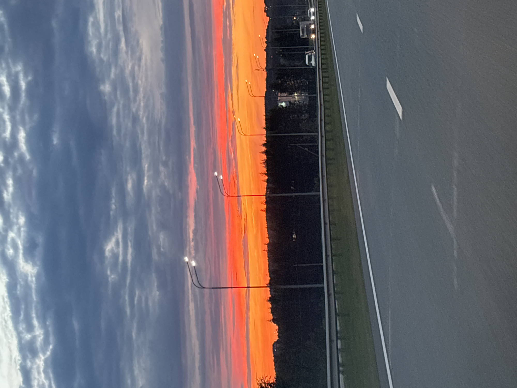 The media file shows a sunset scene along a highway. The sky is ablaze with vibrant hues of orange, red, and pink, transitioning to darker blues and grays as it extends towards the left of the frame. Wispy clouds are visible against the colorful sky. Along the edge of the highway, several lampposts with lit bulbs stand at intervals. A line of trees forms a dark silhouette against the sunset, separating the sky from the road. The highway itself is asphalt, with white lane markings visible. A few vehicles, appearing as headlights and taillights, are traveling on the road, suggesting it is either dusk or early evening. The overall atmosphere is serene and picturesque, capturing the beauty of a sunset over a travel route. The location context indicates this scene is in Bogorodskoe, Russia. No visible text is present in the image.