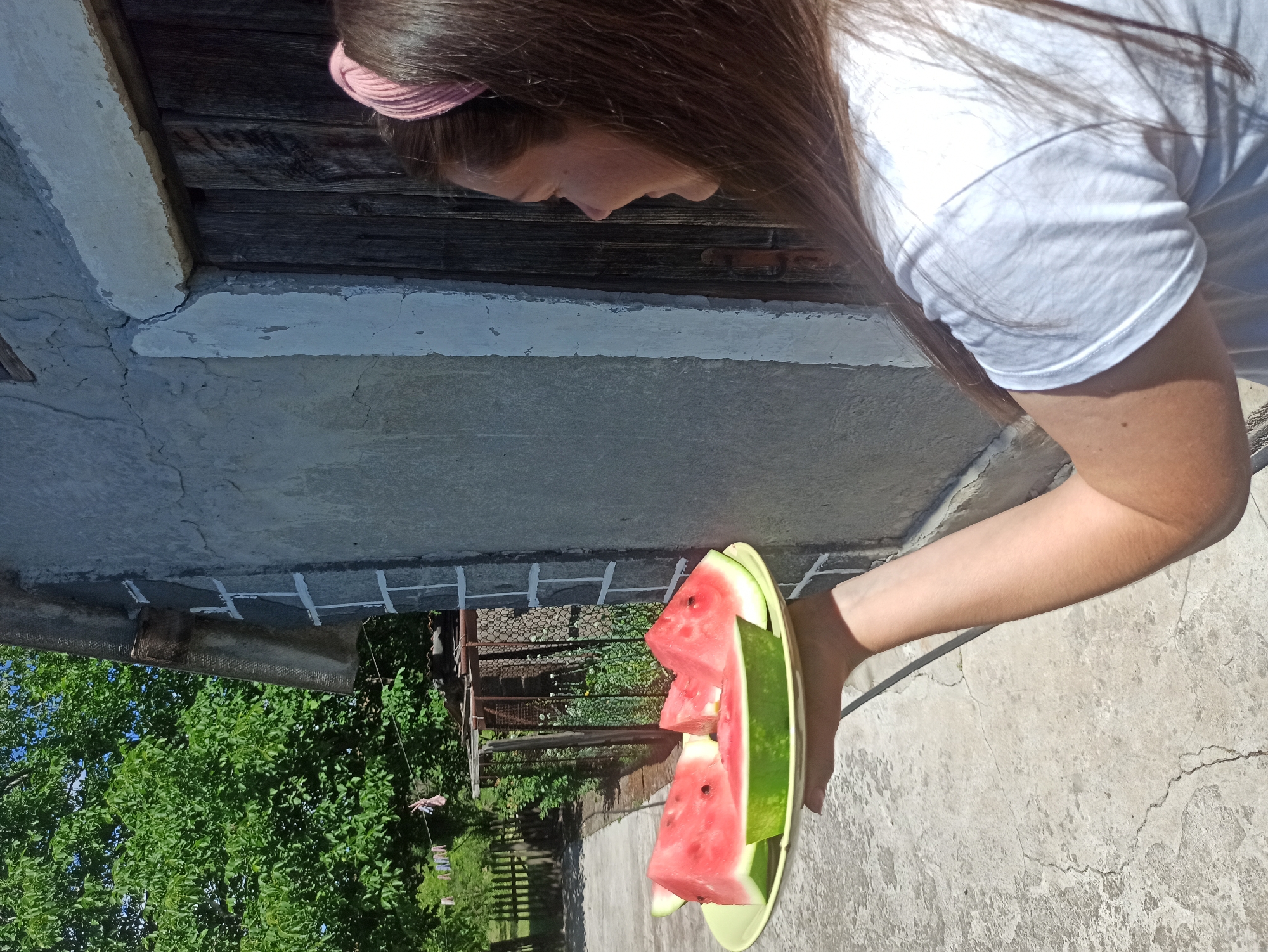 A person holding a plate of watermelon slices stands outdoors, with a rough-textured building to their right and trees and a fence in the background. The person is wearing a white t-shirt and has long brown hair, with a pink scrunchie visible. The watermelon slices are triangular and have visible seeds. The ground is concrete with visible cracks. The sky is blue and clear. The image appears to be taken during the daytime. No specific location in Kryvyi Rih, Ukraine, is identifiable from the visual information.