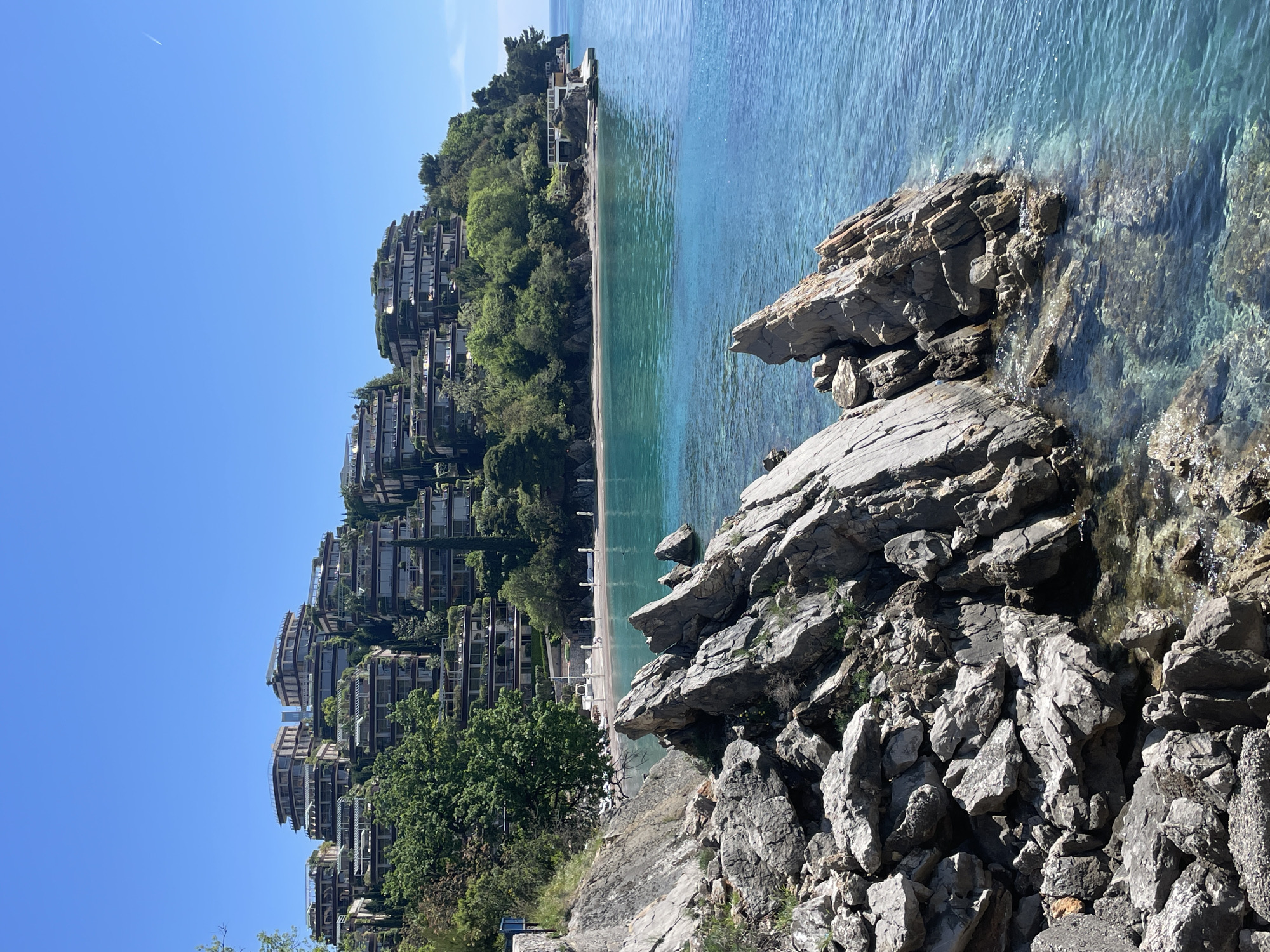 This image captures a vibrant and serene coastal landscape in Budva, Montenegro, under a clear blue sky.
In the foreground, on the right side, large, rugged grey rock formations jut out into exceptionally clear, turquoise-blue water. The water around these rocks is calm, with small ripples and splashes where it meets the stone, revealing the rocky seafloor below.
The midground features a beautiful sandy beach, distinct with its light-colored sand. Several white sun umbrellas and sunbeds are neatly arranged along the shore, suggesting a well-maintained resort or private beach area. The water along the beach is a striking light turquoise, transitioning to a deeper blue further out. At the far end of the beach, another building, possibly a beach club or restaurant, is visible right at the water's edge, complete with a small pier.
The background is dominated by a lush, green hillside densely covered with trees. Integrated into this natural landscape are multiple modern, terraced buildings. These multi-story structures, likely luxury apartments or resort accommodations, feature balconies and appear to be built into the incline, blending with the surrounding greenery.
The sky is a bright, cloudless blue, with a faint white contrail from an airplane visible in the distance, indicating clear weather. The scene is bathed in bright daylight, suggesting a sunny morning or early afternoon. There are no people visible, and no discernible text. The overall impression is one of natural beauty, tranquility, and upscale coastal living.