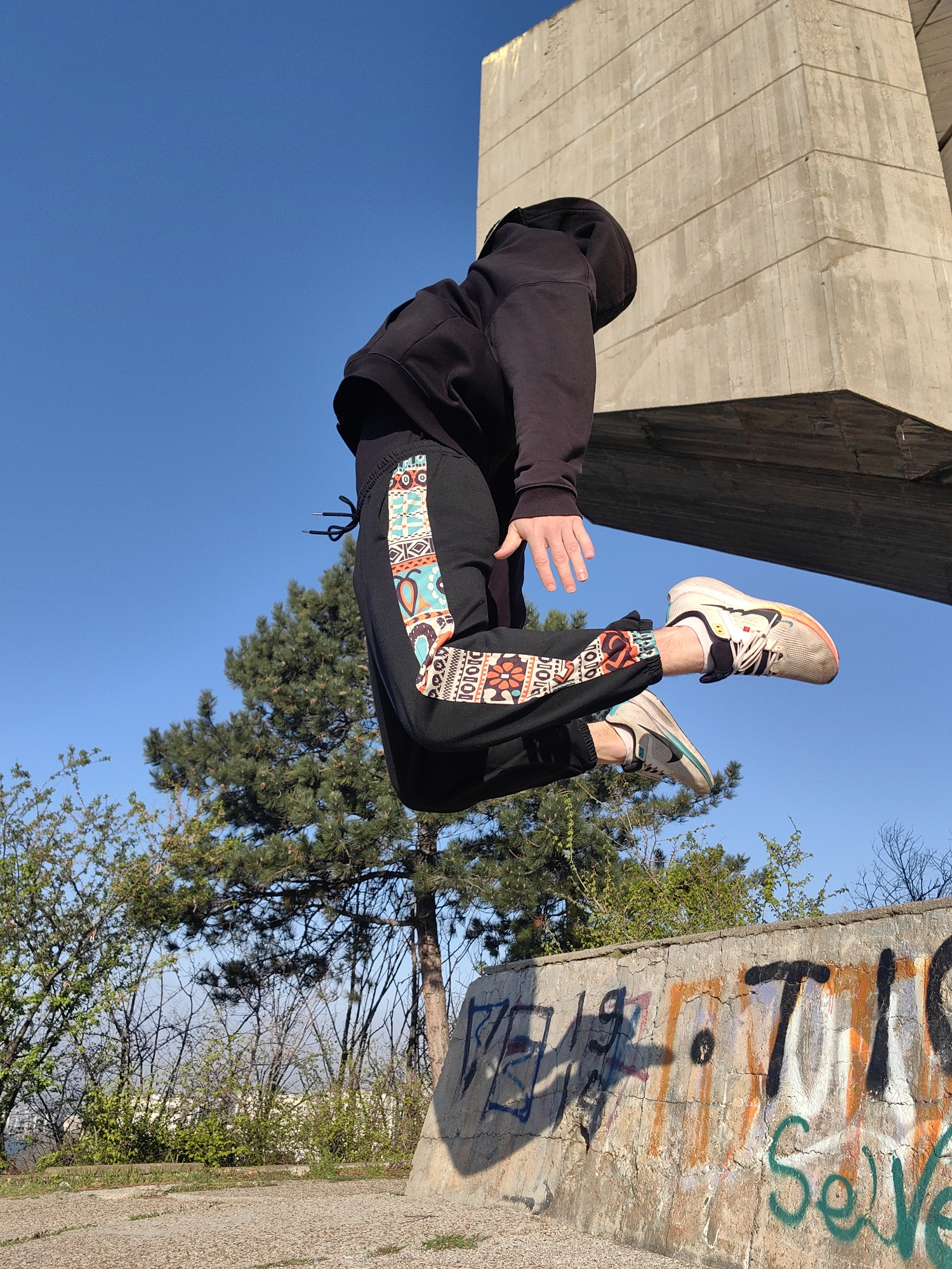 A person wearing a black hooded sweatshirt and black pants with a patterned side stripe is captured mid-air in a jump. The individual's face is obscured by the hood. They are wearing athletic shoes. The backdrop includes a concrete structure with an overhanging ledge, likely part of a monument or building. In the background, trees with green foliage and clear blue sky are visible. A concrete wall with graffiti is in the foreground. Based on the architecture, landscape, and graffiti style, this image likely depicts a scene at the Pantheon monument in Varna, Bulgaria.