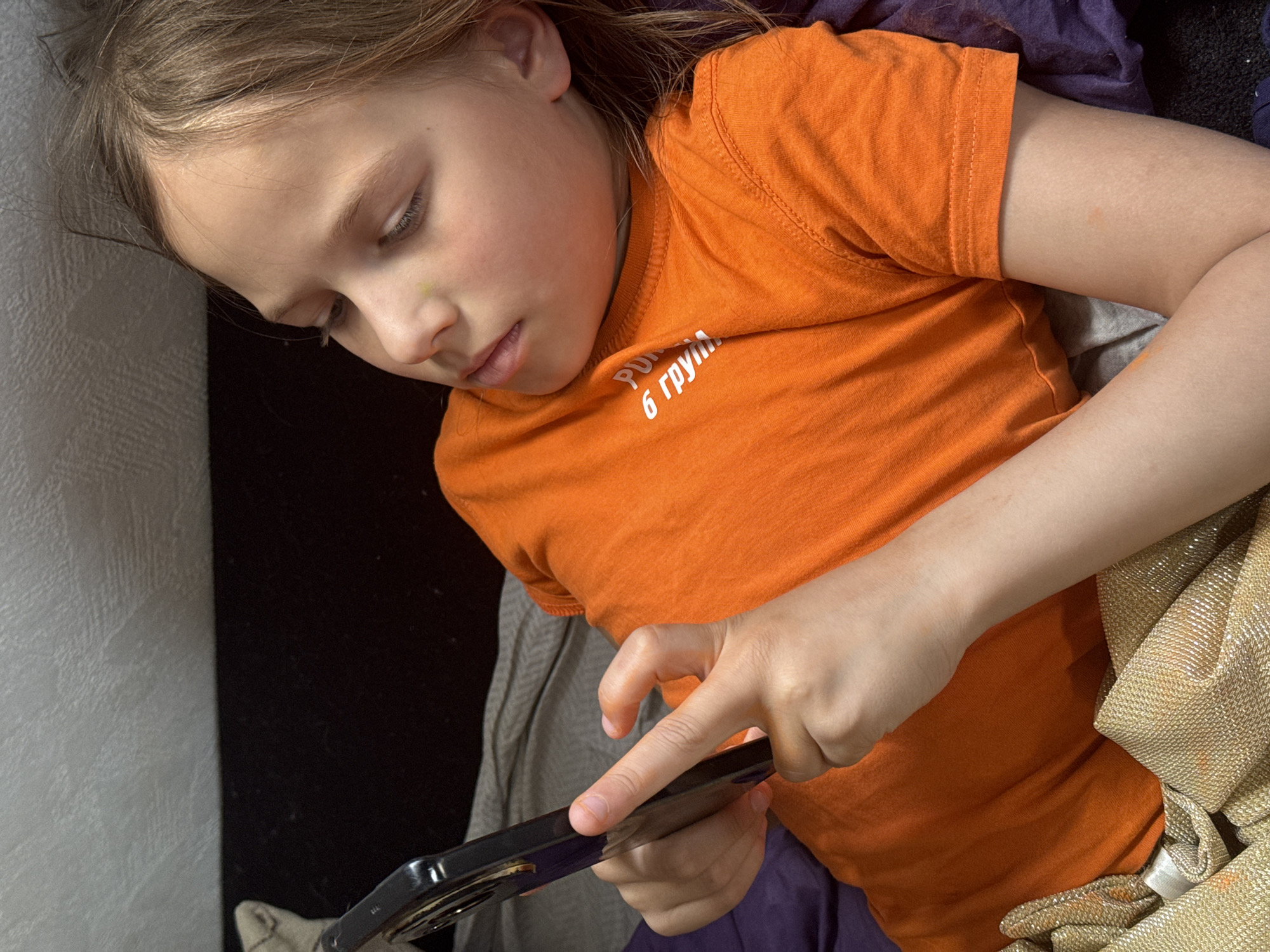 A young girl with light blonde hair is lying down, wearing an orange t-shirt with Russian text on it. She is holding and looking at a black smartphone. The background includes a black and white patterned surface, a dark purple fabric, and a shiny gold-colored cloth. There is no information to conclude that the photo was taken in the city of Rusalevka, Russia.