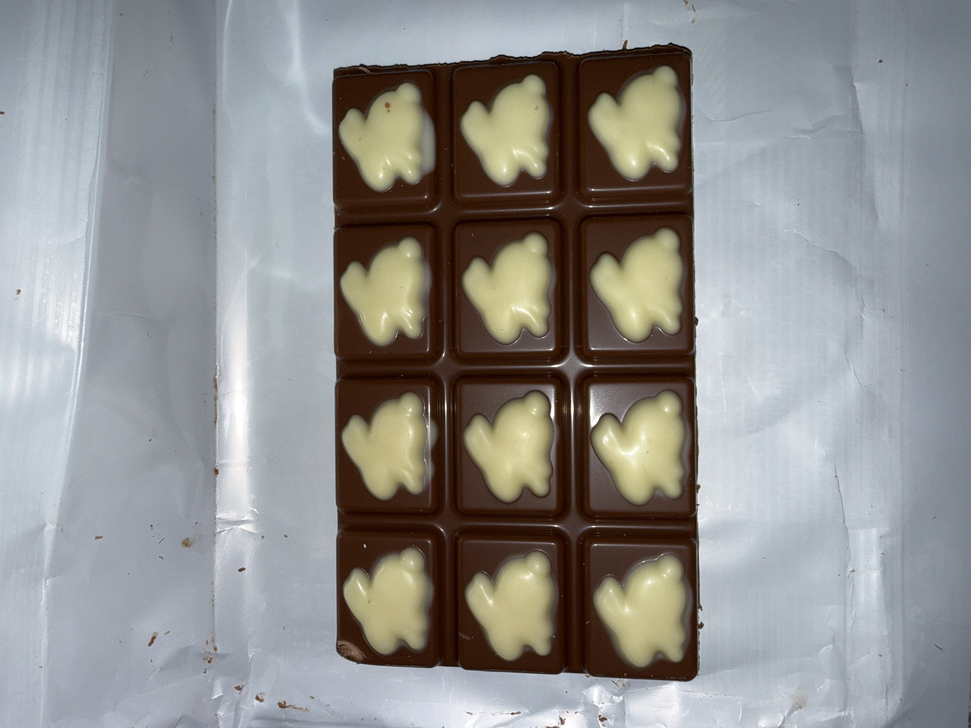 A bar of chocolate lies on a silver, crinkled wrapper. The chocolate is dark brown and divided into twelve square segments, arranged in a 4x3 grid. Each segment is embossed with a white, amorphous shape. The wrapper has some crumbs on it.
The image does not provide any information about the location in the city of Stara Zagora, Bulgaria.
