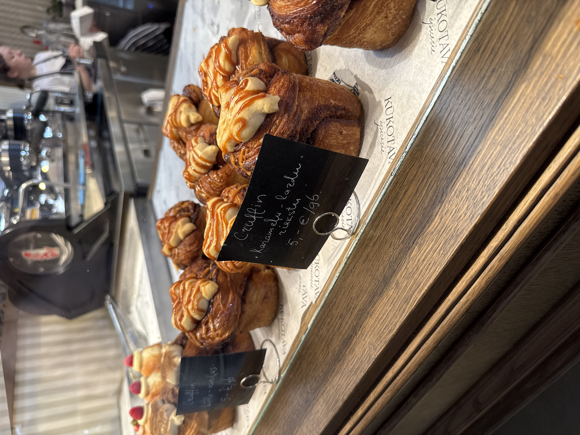 The image shows a display case in a bakery in Riga, Latvia. The case contains several pastries, identified as "Cruffin," a croissant-muffin hybrid. One type is labeled as "Karamelu - lazdu riekstu," indicating caramel and hazelnut flavor, priced at 5.95 euros. Another variety is "Lilly-vanilas," priced at 5.00 euros. The brand "KUKOTAVA épicerie" is visible on the display paper. In the background, there is a partial view of a person in a barista uniform standing near coffee-making equipment. The display case has a wooden frame and a transparent front, allowing the pastries to be viewed.