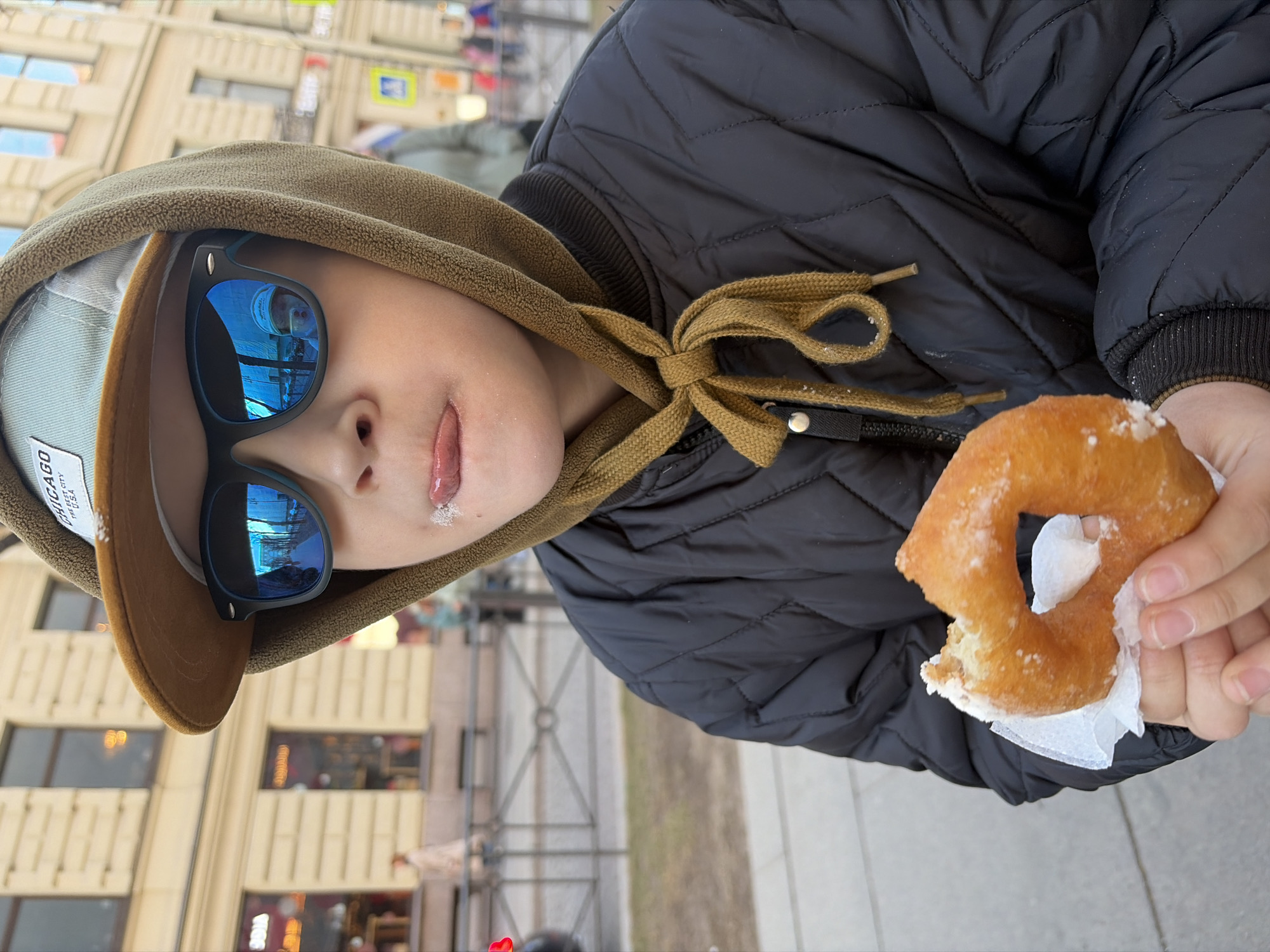 A child is pictured eating a donut on a street in Saint Petersburg, Russia. The child is wearing a black puffy jacket, a hooded sweatshirt, sunglasses, and a baseball cap. The child has sugar on their face from the donut. The donut is in the child's right hand and has a bite taken out of it. In the background, a street and the exteriors of buildings typical of Saint Petersburg can be seen. The reflections in the child's sunglasses show the buildings in the background.