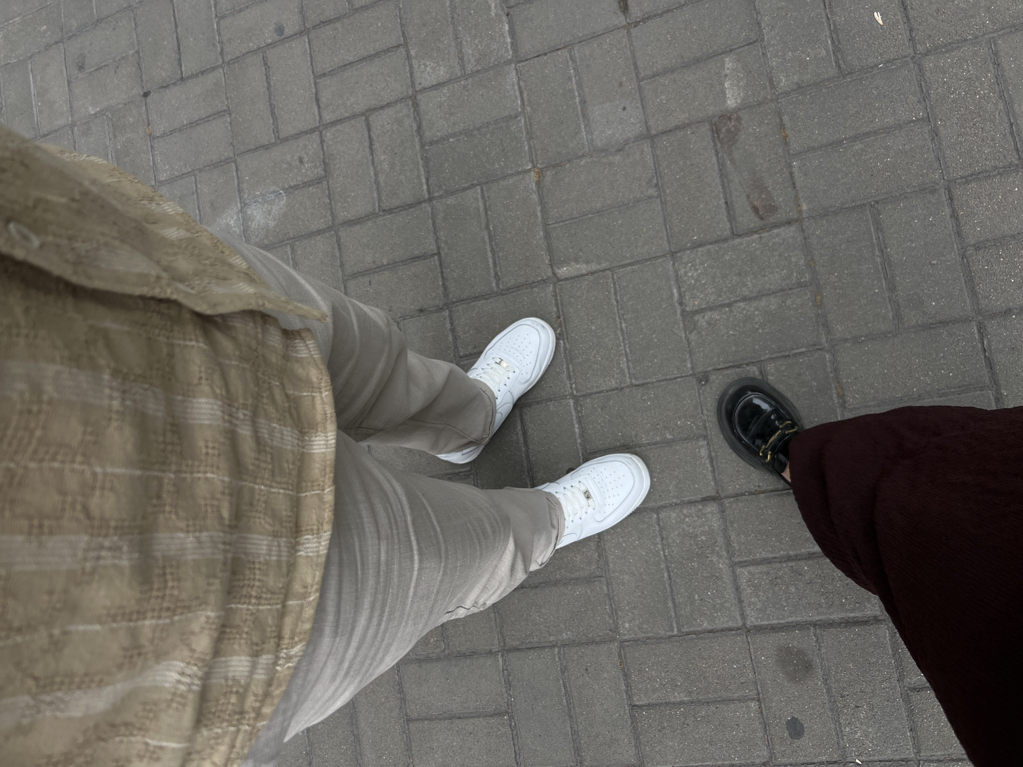 The image shows a view looking down at two pairs of legs and feet standing on a paved area. On the left side, a person is wearing light tan-colored pants and white sneakers. The upper part of their body is partially visible, wearing a patterned beige and white shirt. On the right side, another person is wearing dark brown trousers and black shoes with a shiny finish. The ground is made of gray rectangular pavers. The location is in an urban area of Unknown, Unknown.