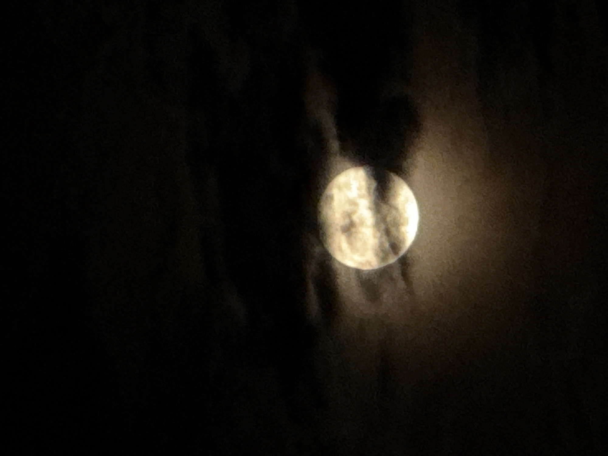 The image shows a full moon partially obscured by clouds. The moon appears bright and textured, with visible craters and surface details. The clouds surrounding the moon are dark and diffuse, creating a dramatic contrast. The overall scene is dark, suggesting a nighttime setting. It is not possible to identify the location in the city of Chaikovsky, Russia, or any people, objects, or interactions from this image.