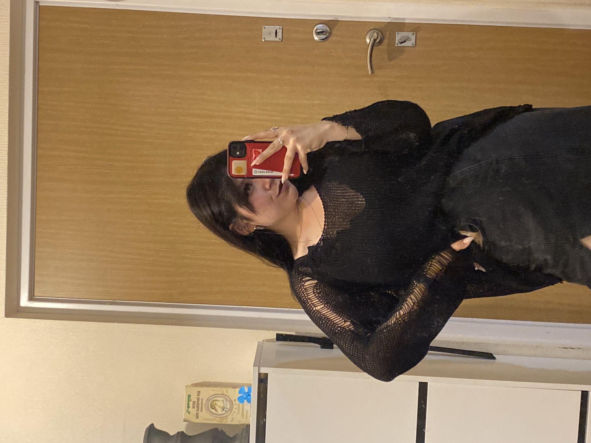 A woman is taking a mirror selfie in an indoor setting, possibly an apartment hallway or entryway, with a wooden door and a white cabinet visible. The woman has dark hair and is wearing a black knit top with cutouts and a dark-colored skirt or shorts. She is holding a red smartphone in her right hand to take the picture. The background includes a white cabinet with items on top, including a box with Cyrillic writing and a blue decorative item. The door has a silver handle and a lock mechanism. There are no indications to suggest this location is in Saint Petersburg, Russia.