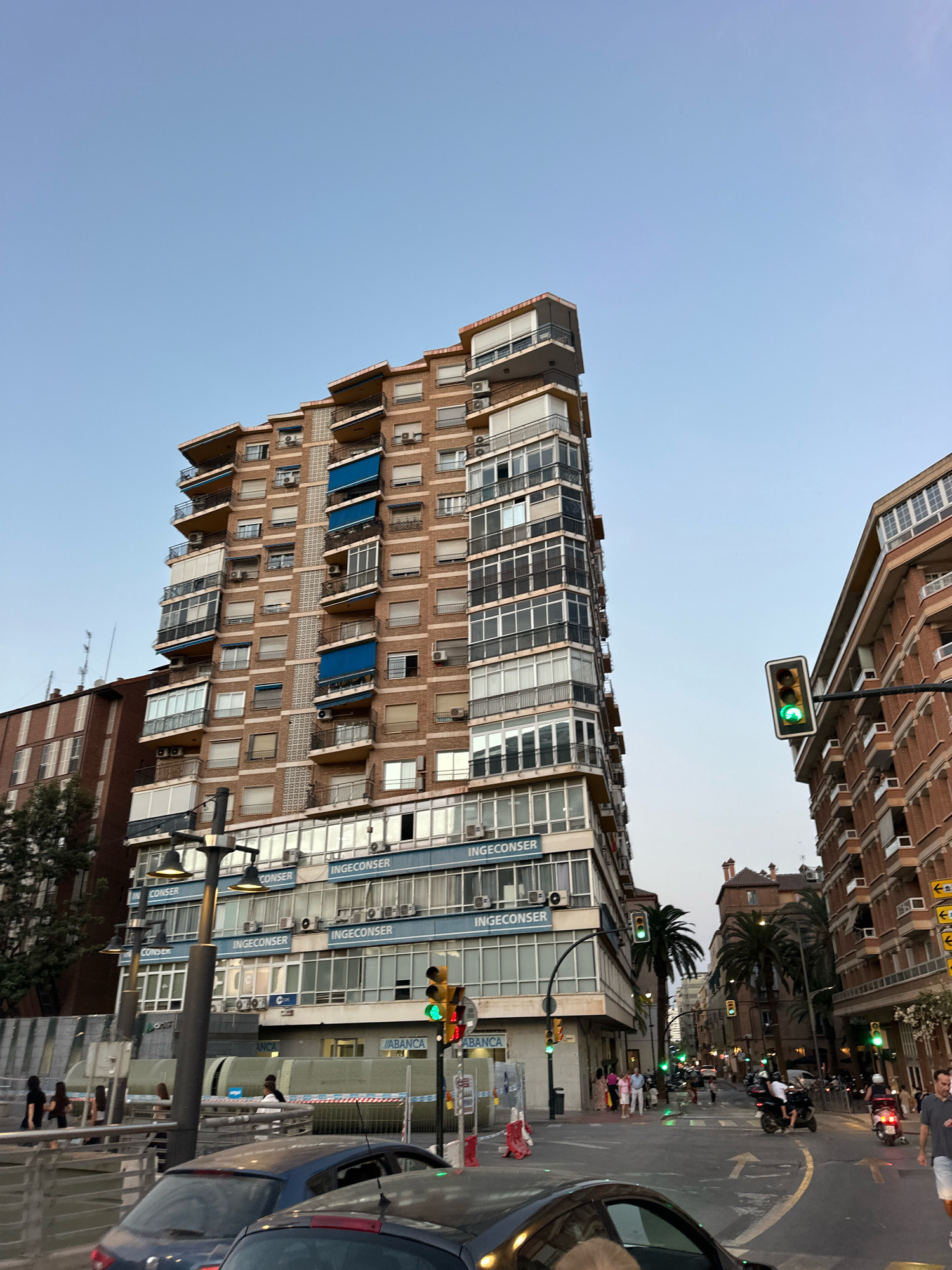 This is a street-level, eye-level shot of a city intersection in Málaga, Spain, likely taken during the late afternoon or early evening. The sky is a clear, pale blue, indicating daylight but with the sun beginning to set.
Dominating the left side of the frame is a tall, multi-story brick apartment building with a distinctive, angular facade that tapers towards the top. It features numerous balconies with dark railings and large windows, many with white roller blinds. Some balconies have blue awnings extended. Air conditioning units are visible on the exterior walls. The lower floors of this building appear to house commercial spaces with large glass windows, displaying signs for "INGECONSER" and "ABANCA".
To the right of the apartment building, another multi-story building constructed with reddish-brown brick is visible. It also has multiple windows and balconies.
In the foreground, two cars are partially visible, one dark blue and another dark grey, suggesting traffic on the street. A street lamp with multiple lights stands prominently in the mid-ground on the left. Traffic lights are active, with a green light illuminated on a signal suspended over the intersection. Several palm trees are scattered throughout the scene, adding a tropical element to the urban environment.
A group of pedestrians is walking on the sidewalk on the left side of the street, near the commercial storefronts. Further down the street, more people are visible, some on scooters or motorcycles, suggesting a lively urban atmosphere. Construction barriers, typically plastic and white with red accents, are present near the sidewalk on the left, indicating ongoing work. The street itself is asphalt, with painted lane markings, including directional arrows. The overall impression is of a typical European city street scene with a mix of residential and commercial architecture, traffic, and pedestrian activity.