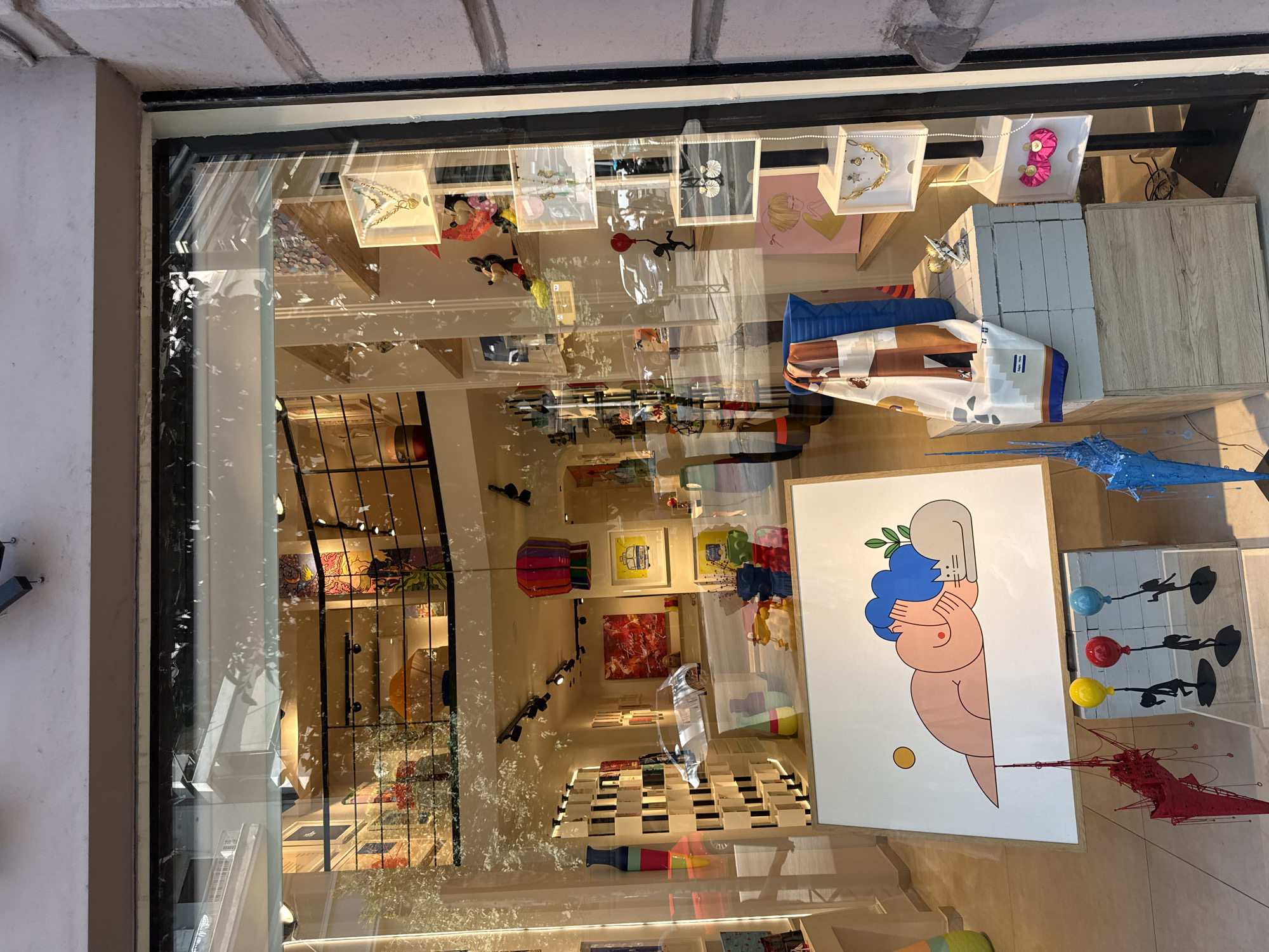 The image shows a street-level view of a shop window display on a sunny day. The shop appears to be an art gallery or boutique, showcasing a variety of items including paintings, sculptures, jewelry, and decorative objects. In the foreground, a framed artwork depicts a stylized nude figure with blue hair, resting with a cat and a yellow sun. Below this artwork, there are several decorative elements including a red, abstract sculpture resembling a crane or machinery, and three silhouetted figures holding balloons.
Further into the display, there are shelves with various items. On the right side, there are several clear acrylic boxes displaying jewelry, including necklaces and earrings. Some Disney figurines, like Mickey Mouse, are also visible on these shelves. In the background, more art is displayed on walls, and a balcony area with railings is visible, suggesting multiple levels within the shop. Reflections on the glass show the street outside, including vehicles and building facades, indicative of an urban setting. The overall impression is a vibrant and eclectic display of art and merchandise.
Based on the style of architecture and the specific items displayed, this shop is likely located in the city of Thessaloniki, Greece, possibly in a commercial or artistic district.