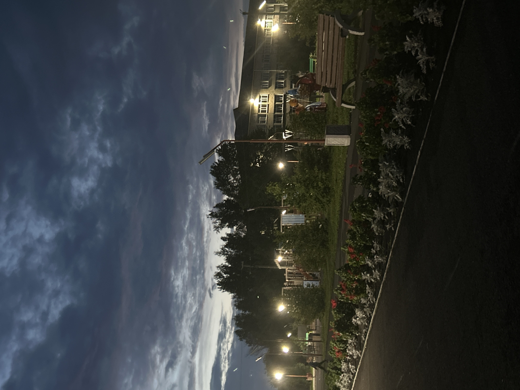 The image captures a nighttime scene in a park or recreational area in Bogotol, Russia. The sky is overcast with dark, textured clouds, suggesting dusk or night. Streetlights illuminate the area, casting a warm glow on the paved walkway and adjacent flowerbeds. A row of red and white flowers lines the walkway, with lush green bushes behind them.
In the background, a multi-story building with visible windows and lights stands under the evening sky, possibly a residential or institutional building. Adjacent to the building is a playground with visible structures, suggesting it's a family-friendly area. There are streetlights scattered throughout the scene, some on tall poles and others integrated into the playground area.
A wooden bench is placed in the foreground, offering a spot for rest. Two individuals are faintly visible in the middle ground; one is walking on the paved path, and another appears to be interacting with playground equipment. The overall atmosphere is calm and serene, illuminated by artificial light against the dark sky.