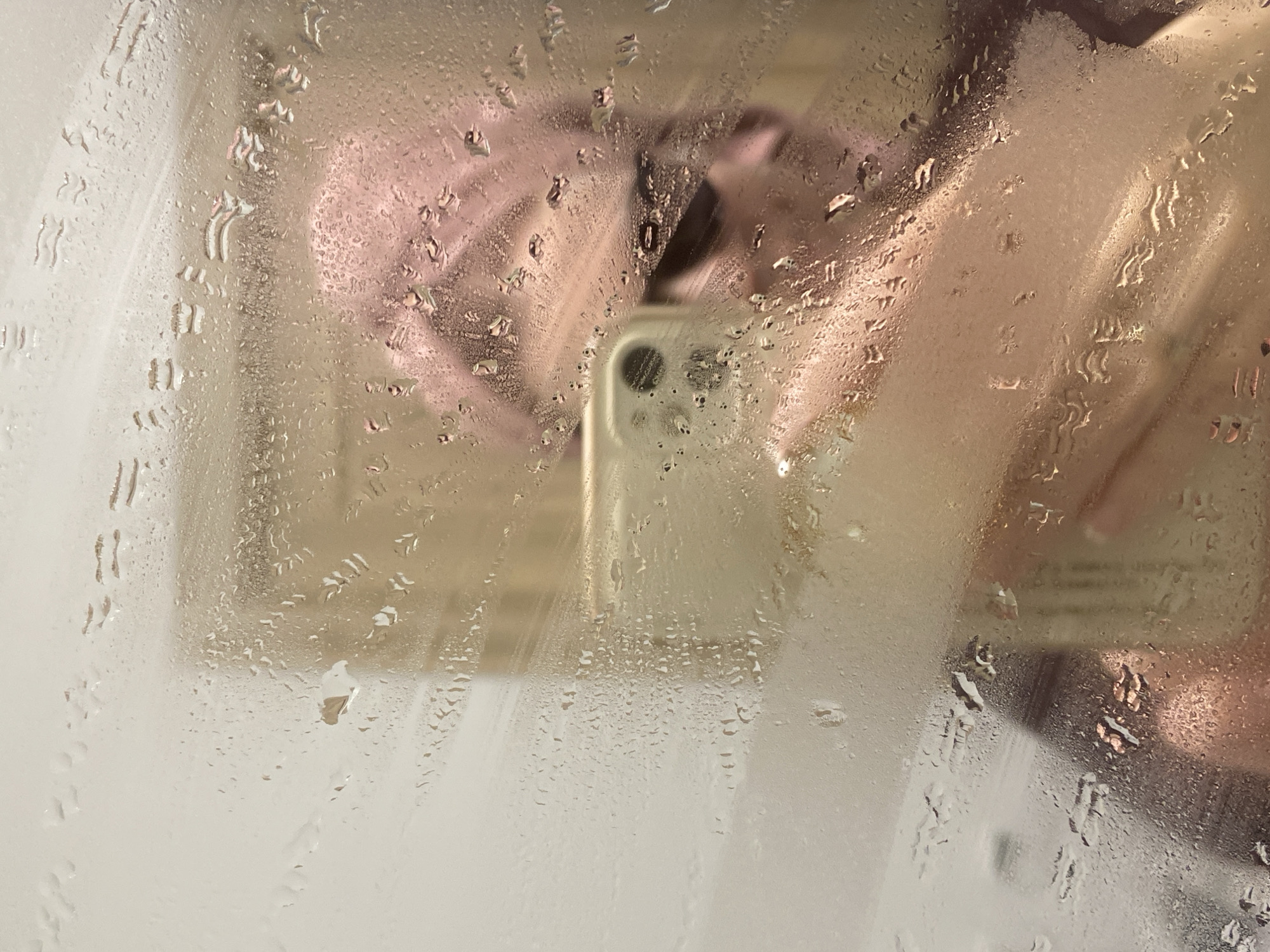 A person holds a smartphone in front of a fogged-up mirror or glass surface covered with condensation. The reflection is blurry, but the person's hands and face are discernible. The smartphone's camera lens is visible. The environment is likely a bathroom or shower stall, given the condensation. There is no discernible evidence to suggest this photo was taken in Yekaterinburg, Russia.