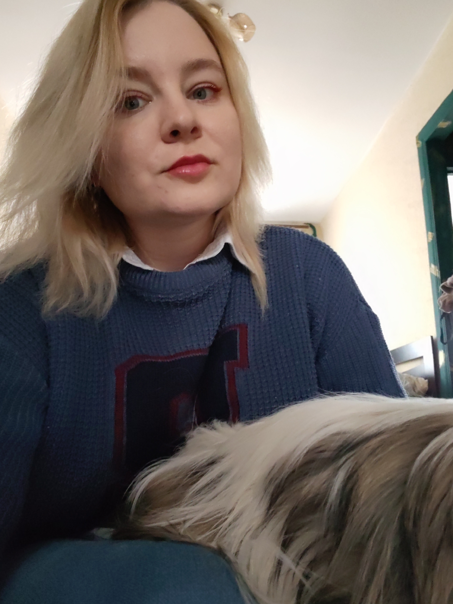 A woman with blonde hair and blue eyes is posing for a selfie indoors. She is wearing a blue sweater with a white collared shirt underneath. Her lips are red. A furry animal, likely a dog, with white and brown fur is partially visible in the foreground. The background shows a cream-colored wall and a doorway with green trim. The location is not discernible as Izhevsk, Russia from the information provided.