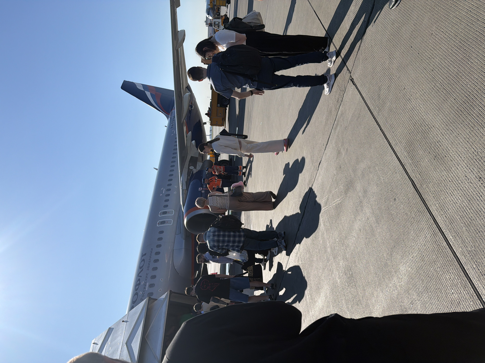 An Aeroflot Airbus A320 aircraft is parked on the tarmac at Sheremetyevo International Airport in Khimki, Russia, under a clear blue sky. Passengers, carrying luggage, are boarding the aircraft via an airstair. The scene is set on a concrete surface marked with lines. The foreground shows the legs and shadows of individuals waiting to board, while the midground features a group of passengers and ground crew in high-visibility vests. The aircraft's livery is visible, with the Aeroflot logo and "РОССИЙСКИЕ АВИАЛИНИИ" (Russian Airlines) text prominently displayed. The tail fin of the aircraft features the Russian flag colors. Other airport vehicles, including a baggage tug, are visible in the background. The lighting suggests it is a sunny day.