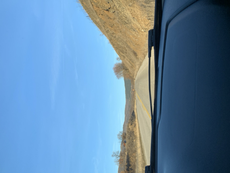 The image presents a driver's-eye view from inside a vehicle, looking out onto a road winding through a dry, hilly landscape in Grevena, Greece. The perspective is through the car's windshield, with the dark dashboard and a segment of a windshield wiper visible on the right side of the frame.
A paved road, marked by a solid yellow double line down its center, curves gently into the distance, suggesting a path through mountainous or rural terrain. On both sides of the road, the land rises into steep, dry hills. These hills are covered with sparse, light brown vegetation and scattered, bare deciduous trees, indicating a dry season, possibly late autumn or winter. The overall color palette of the landscape is dominated by earthy tones of brown, yellow, and orange.
Above the rugged terrain, the sky is a bright, clear blue, stretching across the upper left quadrant of the image. A few faint, wispy cloud formations or contrails are visible, but the sky is predominantly clear and unblemished, suggesting sunny weather conditions. The bright lighting indicates it is daytime, likely morning or early afternoon.
No individuals are visible in the scene, conveying a sense of serene solitude on the road. There is no discernible text within the image. The photograph captures a tranquil moment of travel through a natural, rugged environment.
