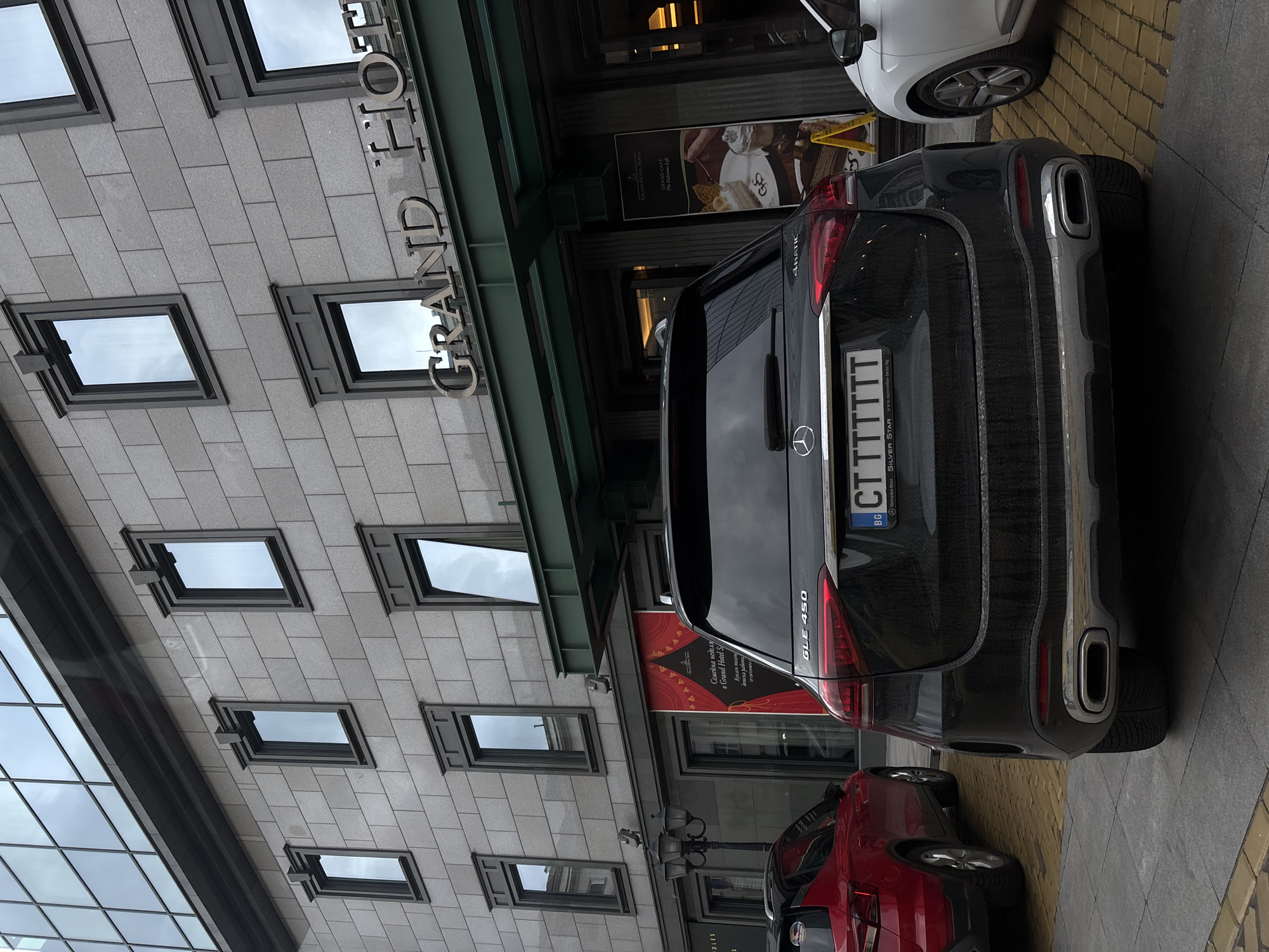 The image shows the rear of a black Mercedes-Benz GLE 450 parked in front of the Grand Hotel Sofia in Sofia, Bulgaria. The car has a Bulgarian license plate that starts with "CT". The building facade is grey with several windows. A red car is parked behind the Mercedes. There is also a white vehicle visible further in front of the Mercedes. A sign for the "GRAND HOTEL SORIA" is visible to the right of the car.