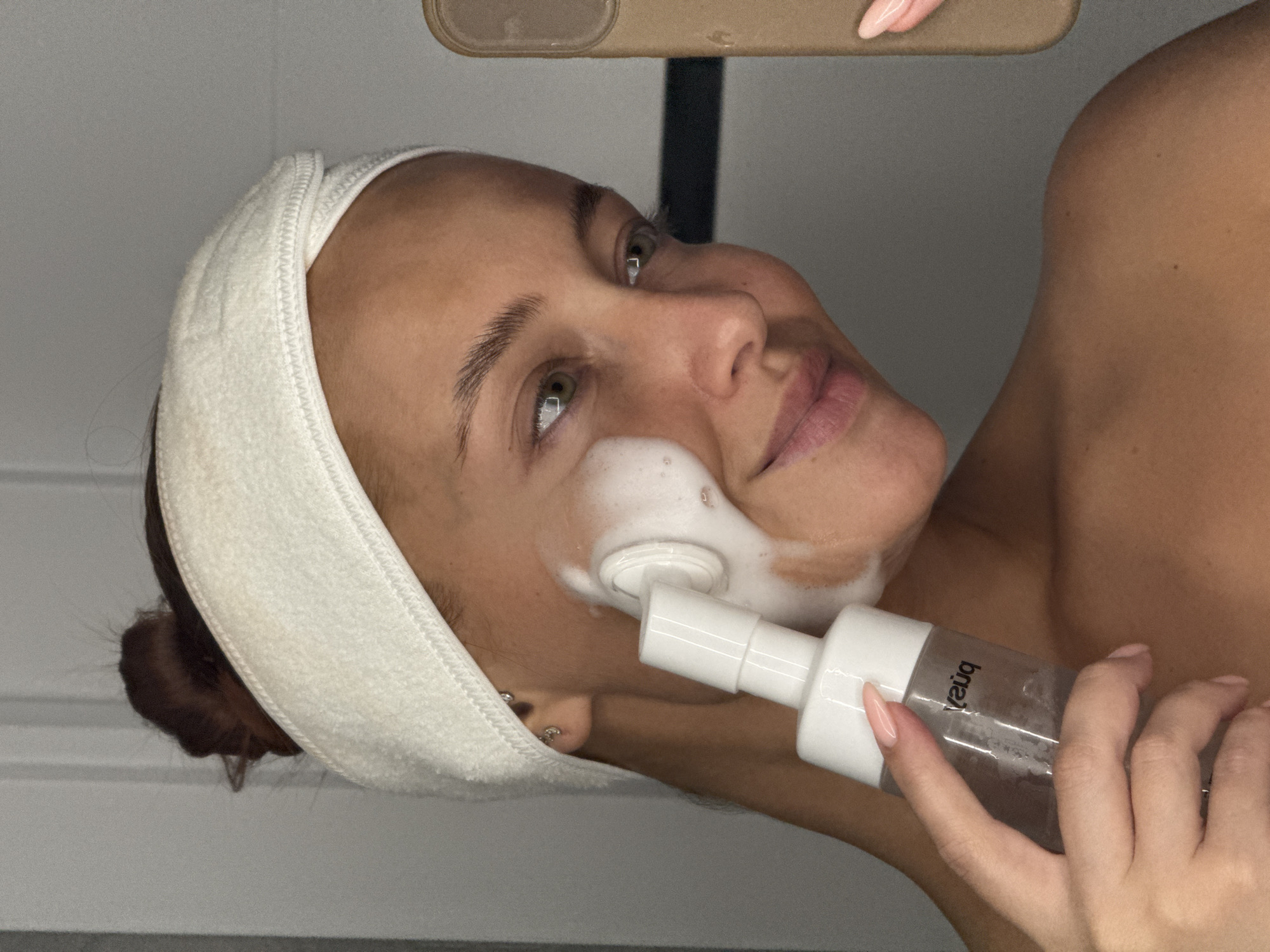 A woman with light skin, reddish-brown hair pulled back in a bun, and a white terrycloth headband is holding a clear plastic bottle with a white pump applicator. The bottle is labeled "brea" in reverse. She has light pink nail polish on her fingernails. There is a white, foamy substance on her face near the applicator. The background is a neutral color, and there is a dark vertical element visible in the upper part of the frame. The location in the city of Chaikovsky, Russia, cannot be determined from the image alone.
