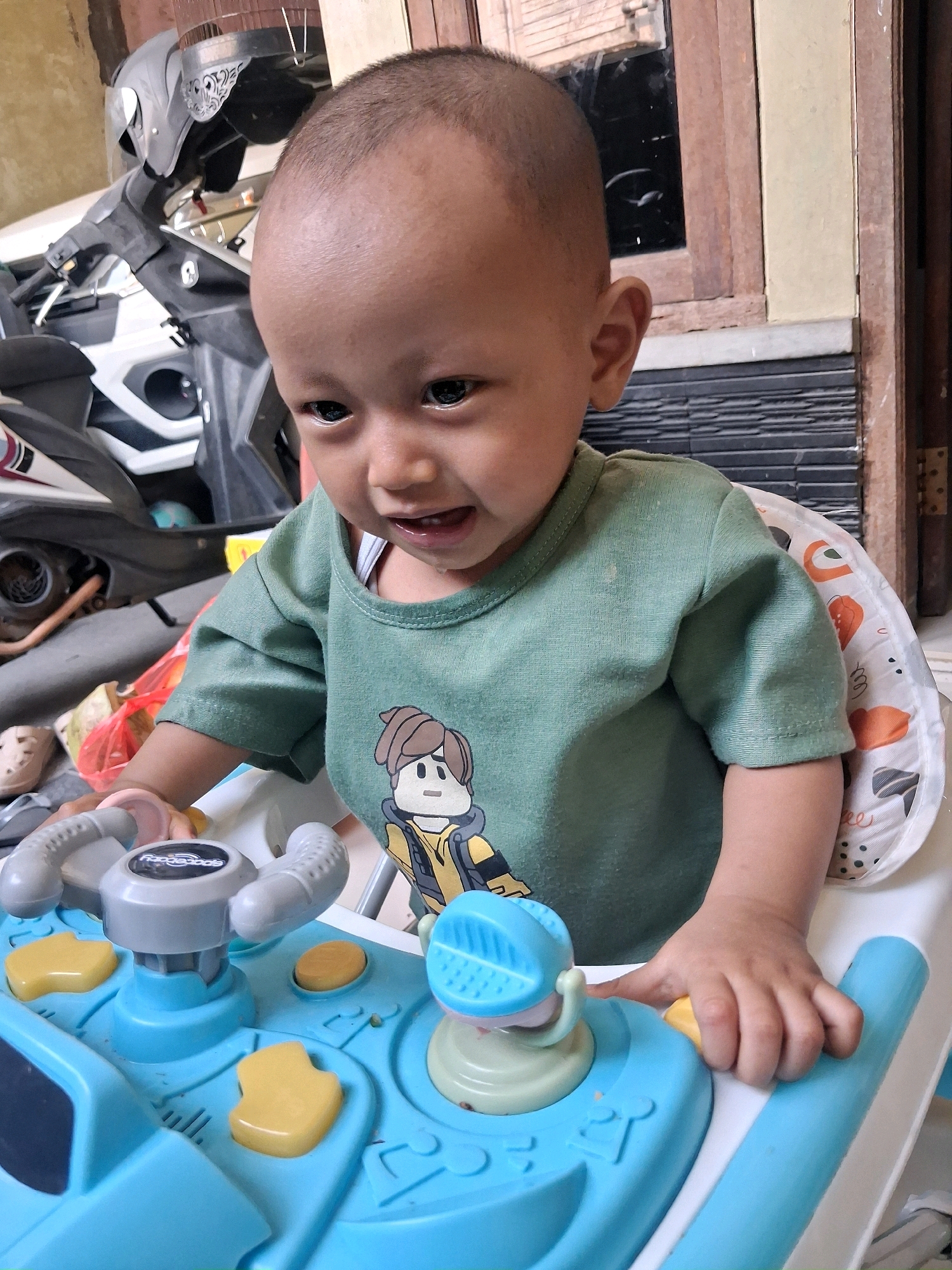 A close-up, eye-level shot captures a young child, likely a toddler, seated in a blue and white baby walker with a playful, patterned seat. The child, with a shaved head and dark eyes, looks slightly to their left, mouth slightly open as if speaking or about to speak. They wear a short-sleeved green t-shirt with a cartoon graphic of a character with brown hair, a yellow jacket, and a grey collar. The child's hands are on the baby walker's blue and grey dashboard, which features various colorful buttons and interactive elements. The background is slightly blurred, revealing a motorcycle parked to the left, suggesting an outdoor or semi-outdoor setting, possibly a home's porch or carport, common in areas like Setu, Indonesia. To the right, a wooden window frame and a section of a wall are visible, further contextualizing the environment.